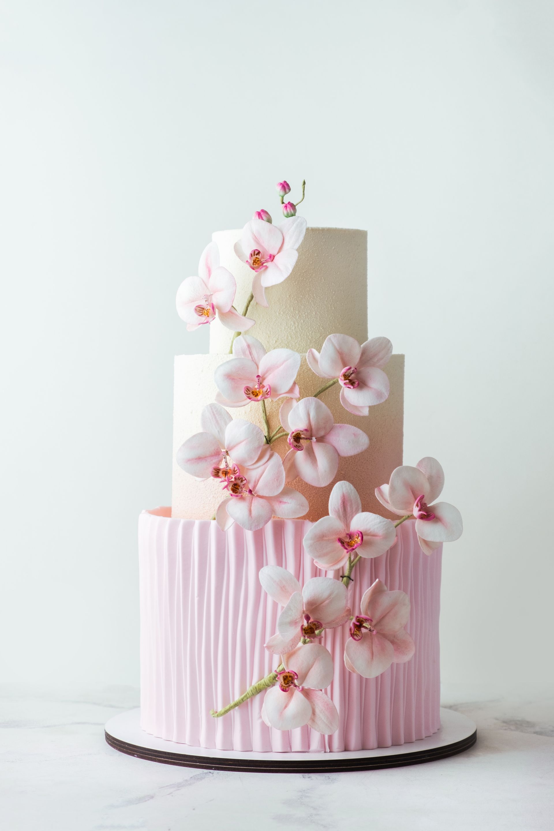 Torta a tre piani con glassa rosa testurizzata, decorata con orchidee rosa, su sfondo bianco.