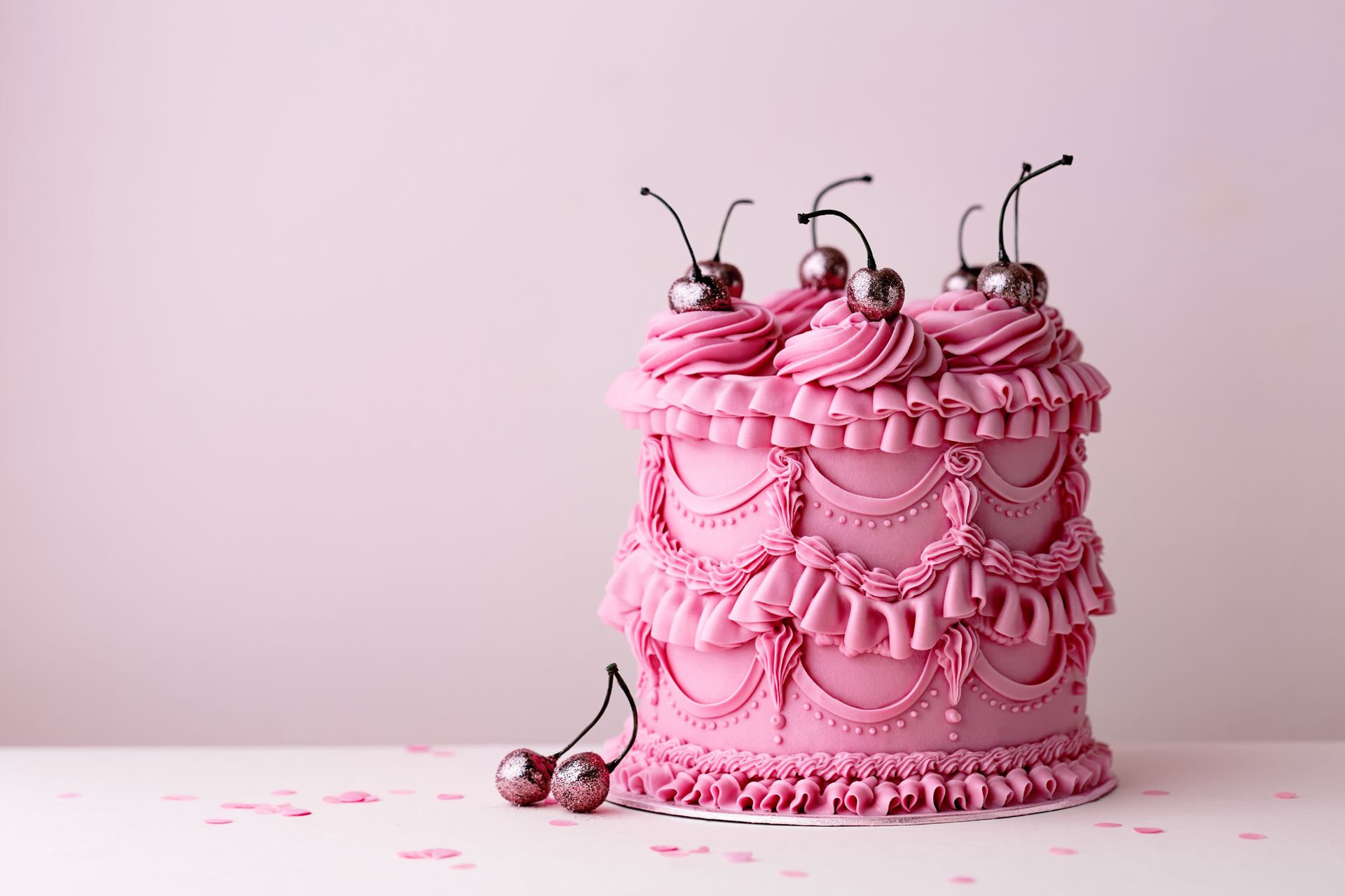 Torta rosa a più piani decorata con volant, perle e ciliegie, su sfondo rosa.