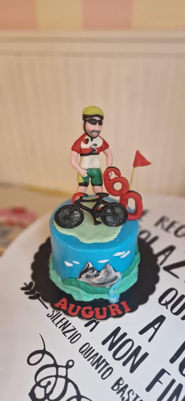 Torta decorata con una statuina di motociclista, montagne e il numero 60, con la parola italiana
