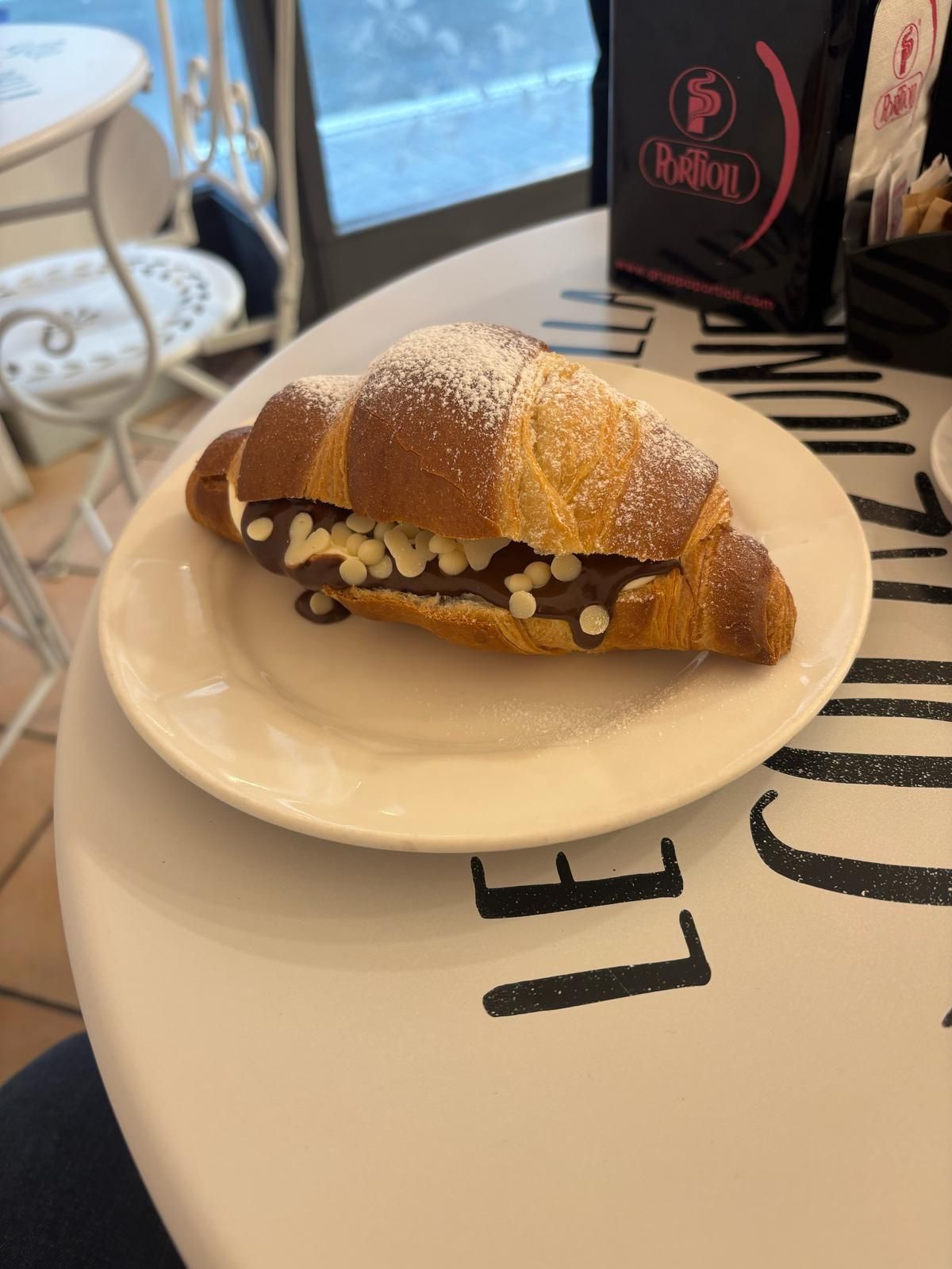 Cornetto farcito con gocce di cioccolato bianco e spolverato con zucchero a velo, disposto su un piatto, sul tavolino di un bar.