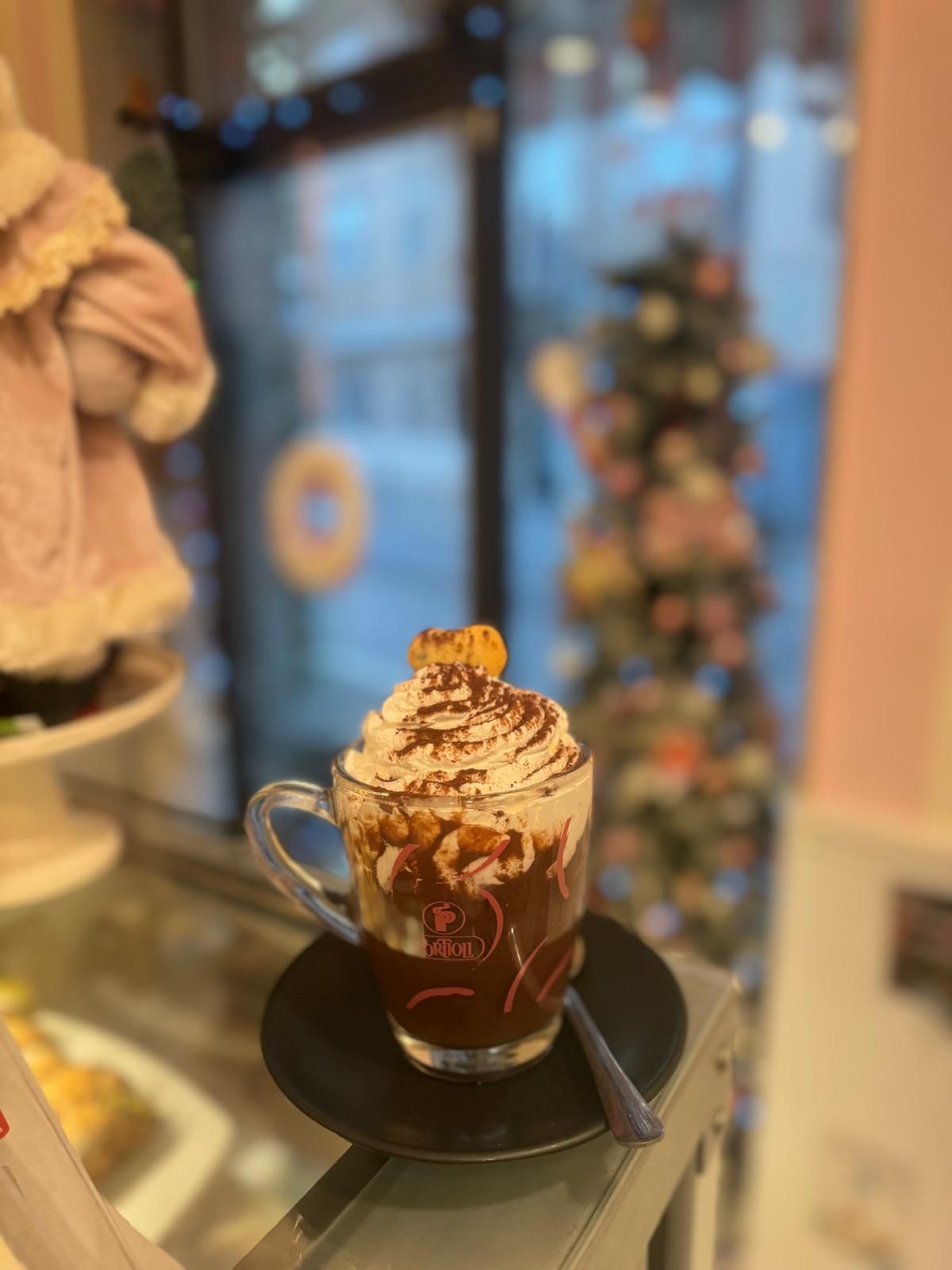 Cioccolata calda con panna montata e caramello, servita su un piattino, in un ambiente da bar con un albero di Natale.