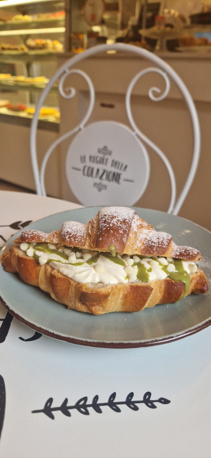 Croissant con ripieno bianco e glassa verde su un piatto blu, accanto a una sedia in un bar.
