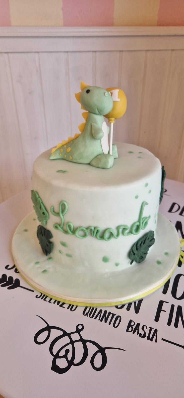 Torta decorata con un dinosauro verde che tiene in mano un palloncino; sul lato è scritto il nome "Leonardo".