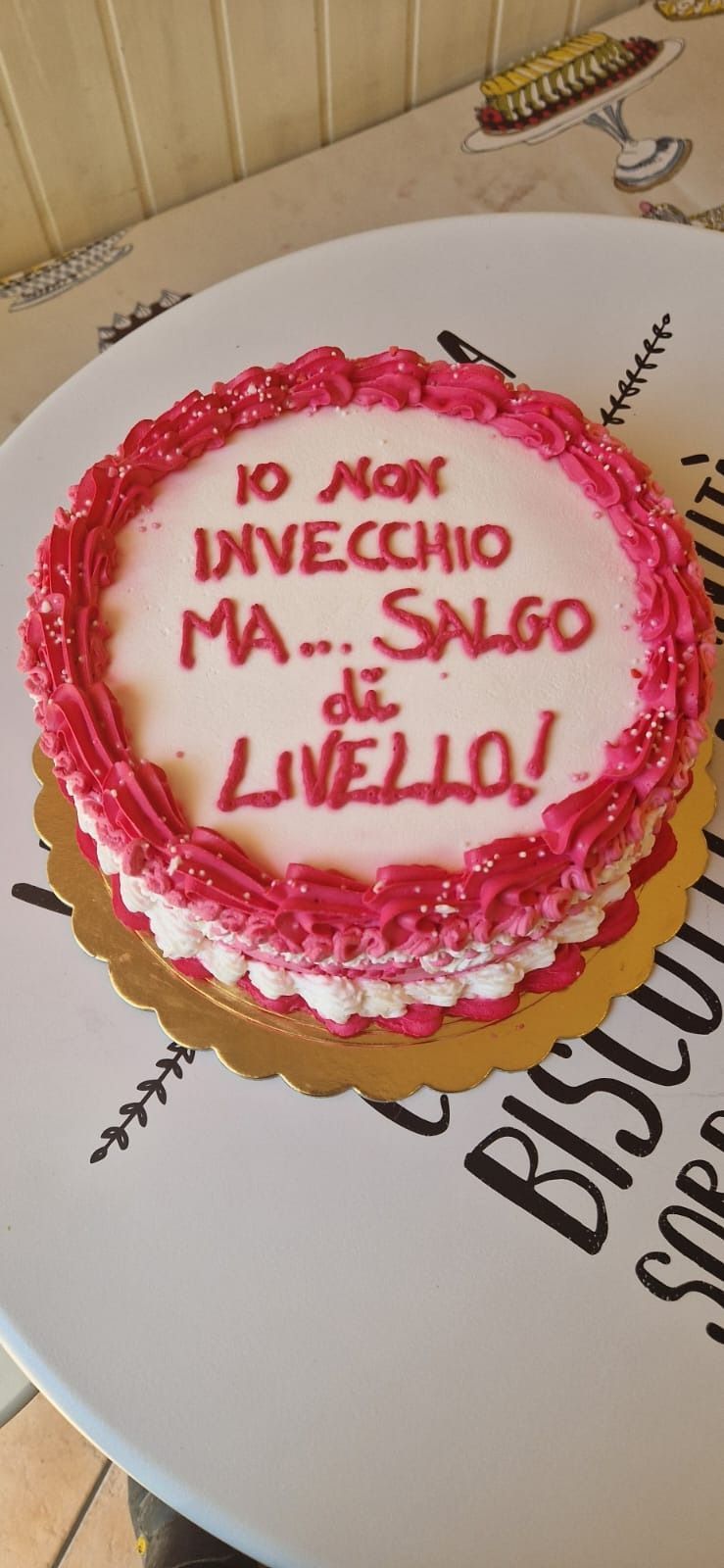 A round cake with pink frosting, it says "Io non invecchio ma... salgo di livello!" on top.