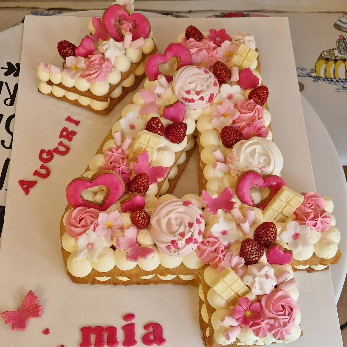 Torta numero 4 decorata con glassa rosa e bianca, fiori e bacche; sul vassoio della torta è scritto