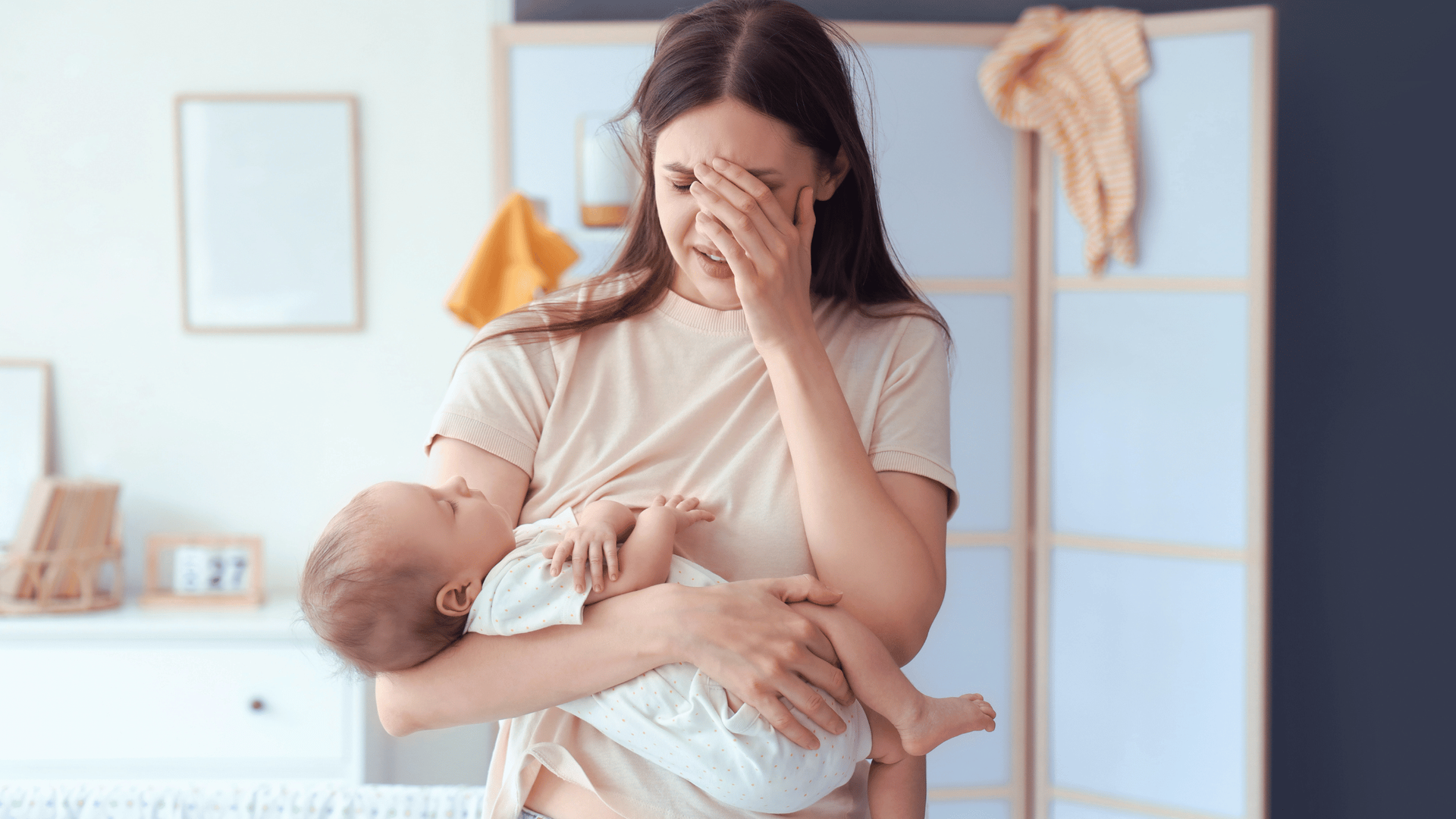 A tired woman holds a sleeping baby in her arms, rubbing her eyes in exhaustion.