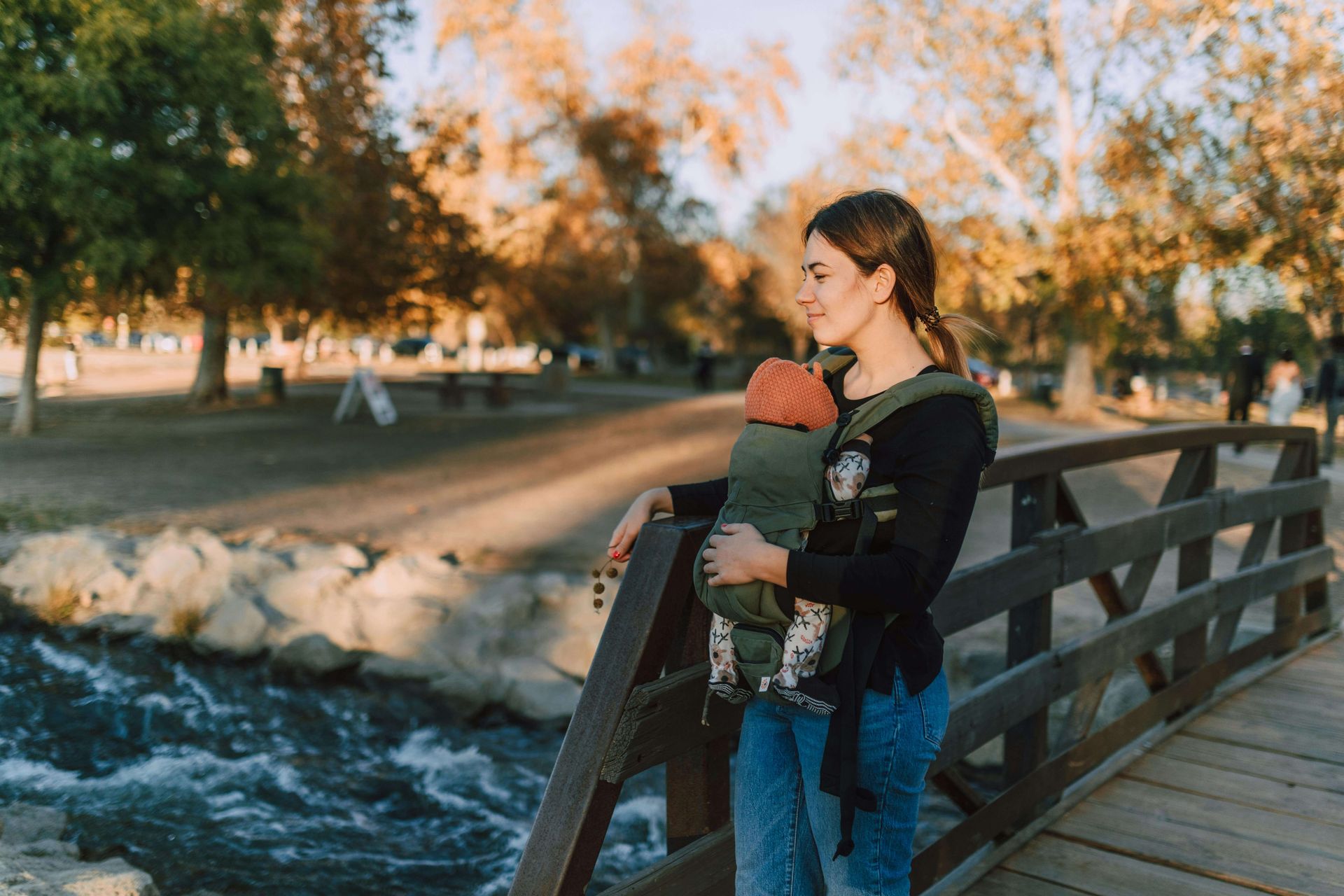 A woman stands on a wooden bridge holding a baby in a carrier, both gazing at the serene river.