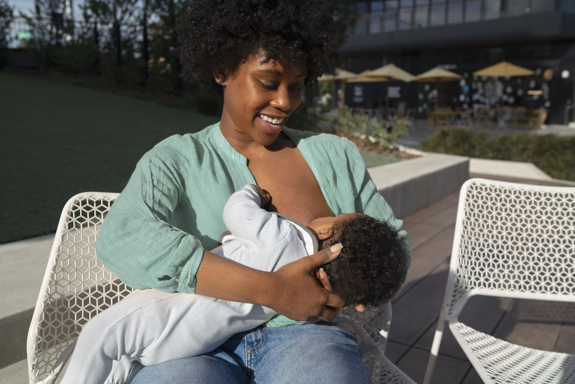 Smiling woman breastfeeding an infant outside on a sunny day, sitting on a white chair.