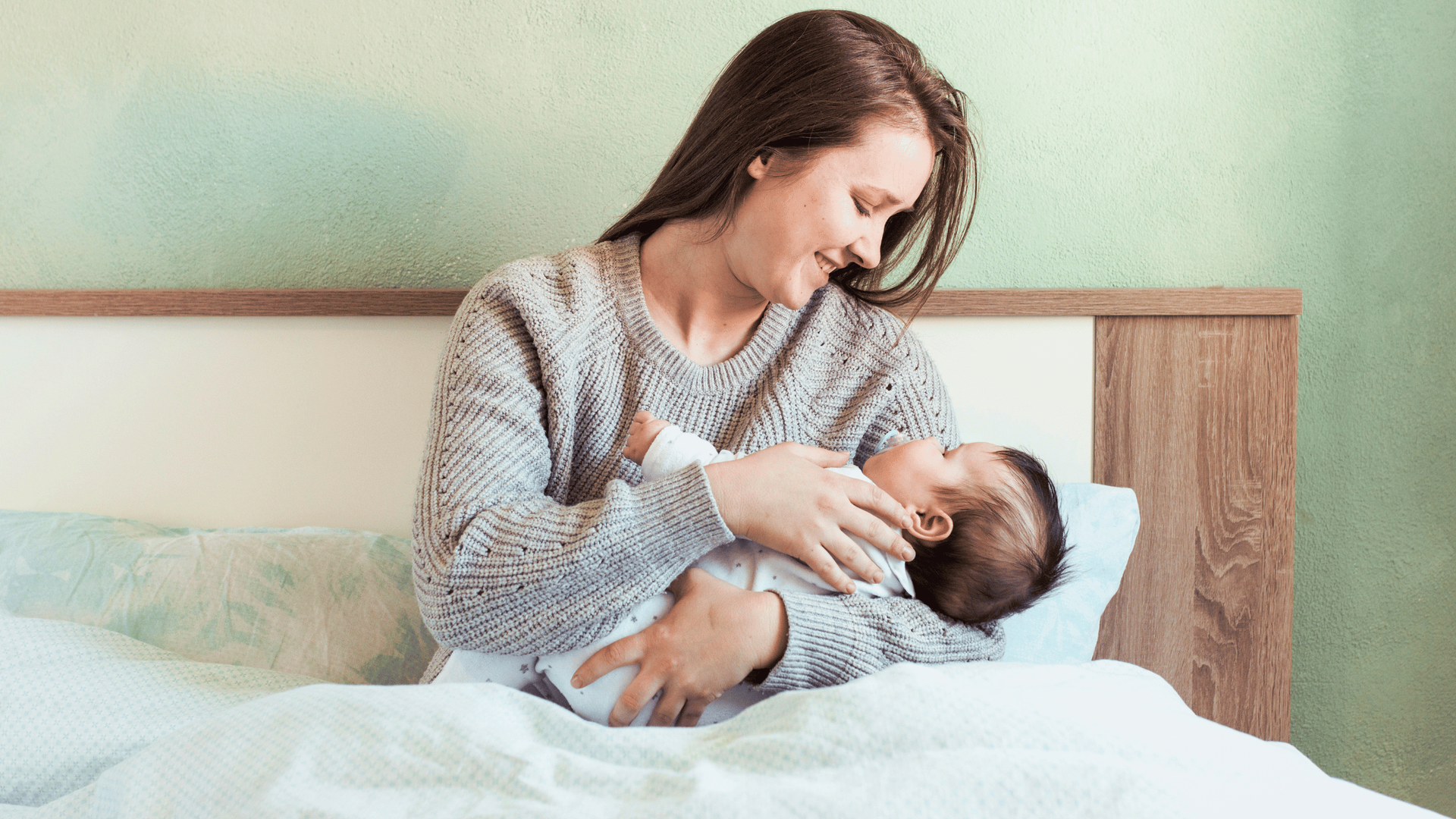 A woman sits on a bed, cradling her baby in her arms, both appearing calm and content.