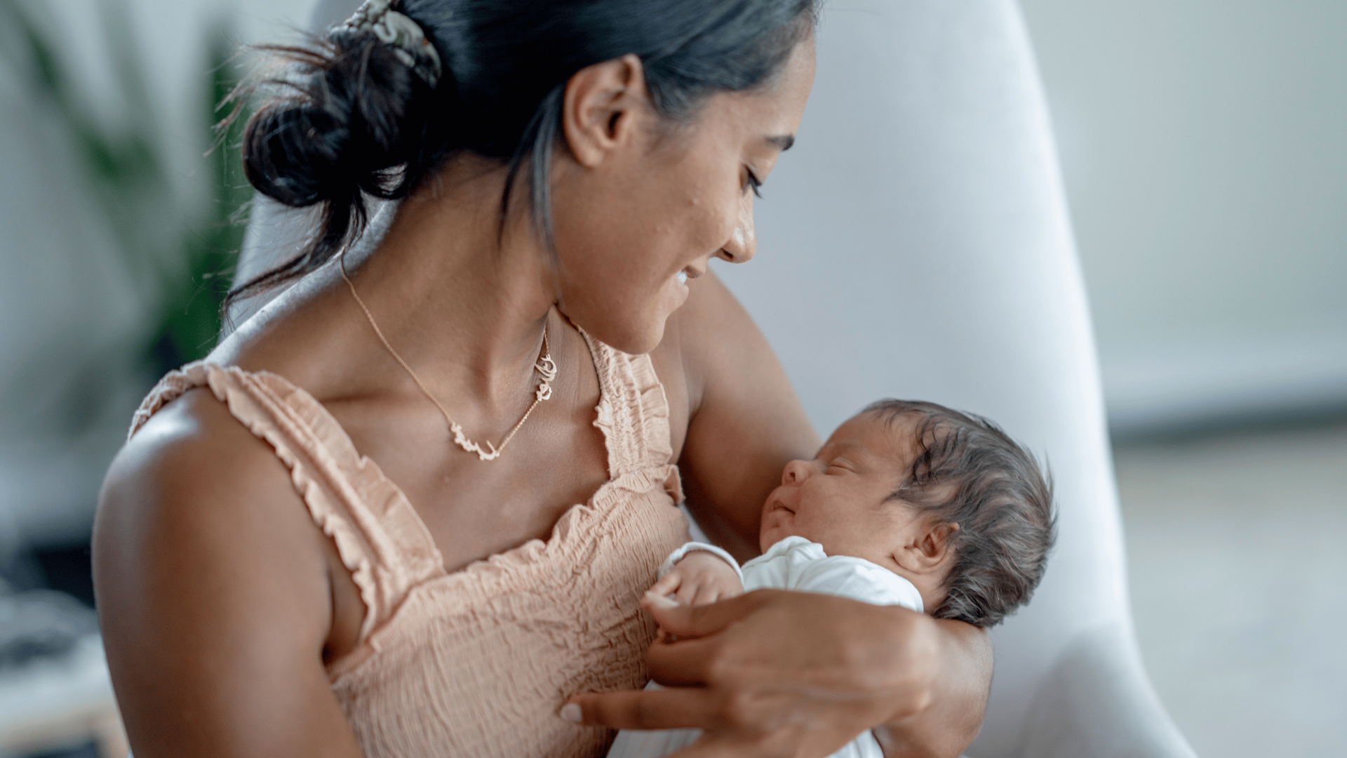 A woman smiles warmly while holding a sleeping baby. She wears a peach-colored top and necklace.