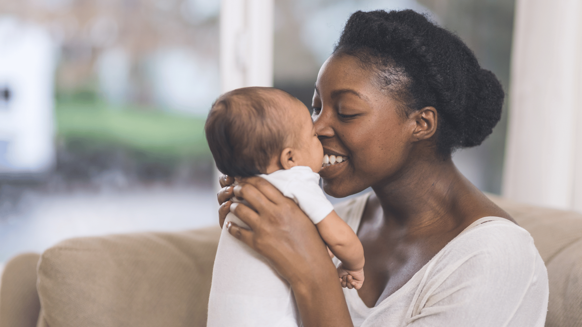 A woman lovingly cradles and smiles at a baby, their faces close. They are indoors, sitting on a sof