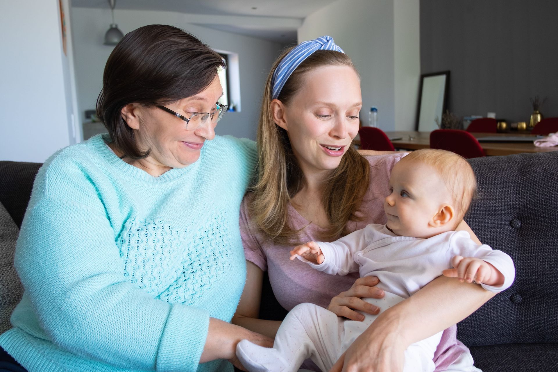 A  woman and an older woman sit on a couch, smiling at a baby on the woman's lap.