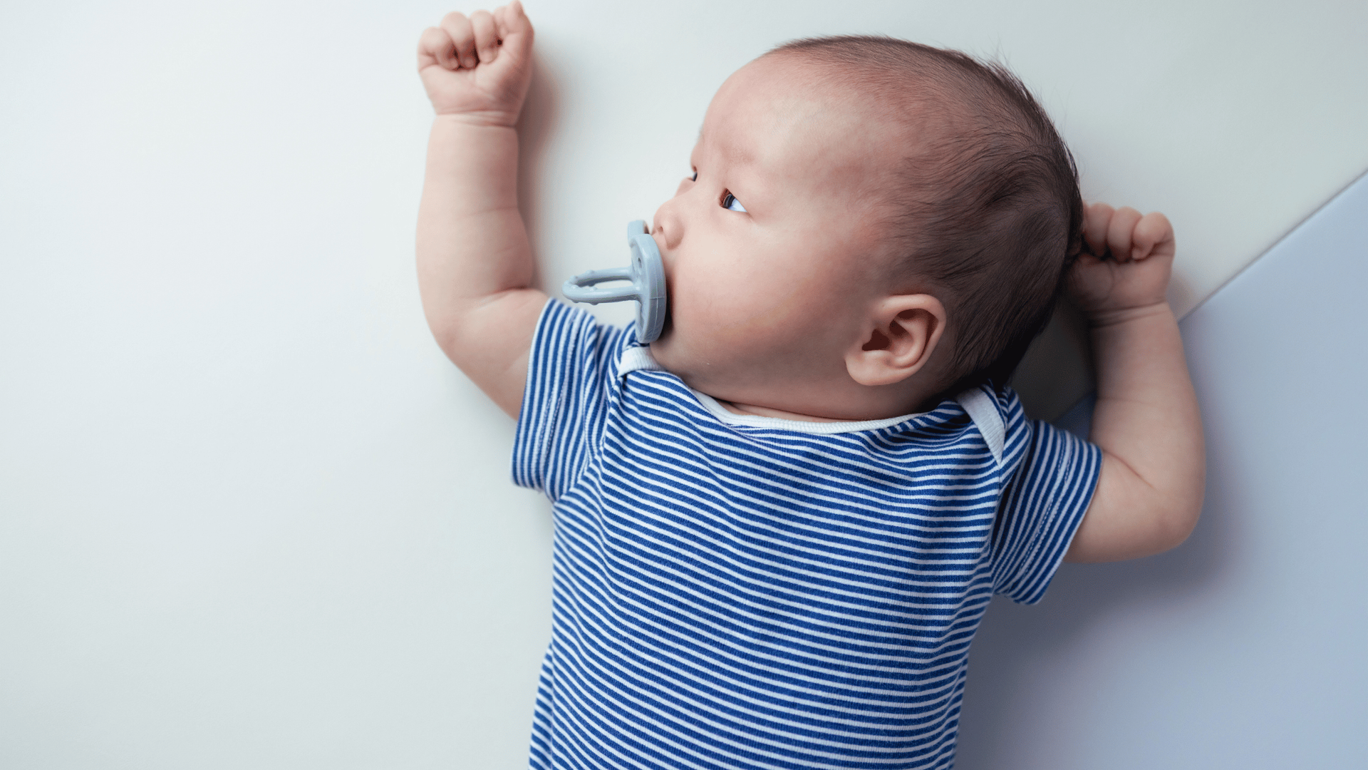 Baby in a blue striped onesie lies on a white surface, gazing upward with a pacifier in mouth.