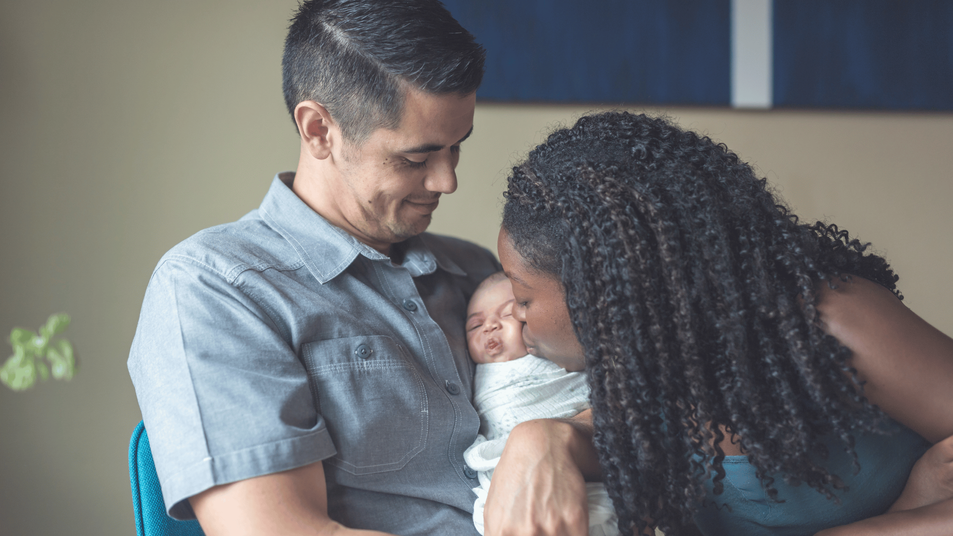 A couple lovingly cradles a sleeping newborn. The mother gently kisses the baby's head.