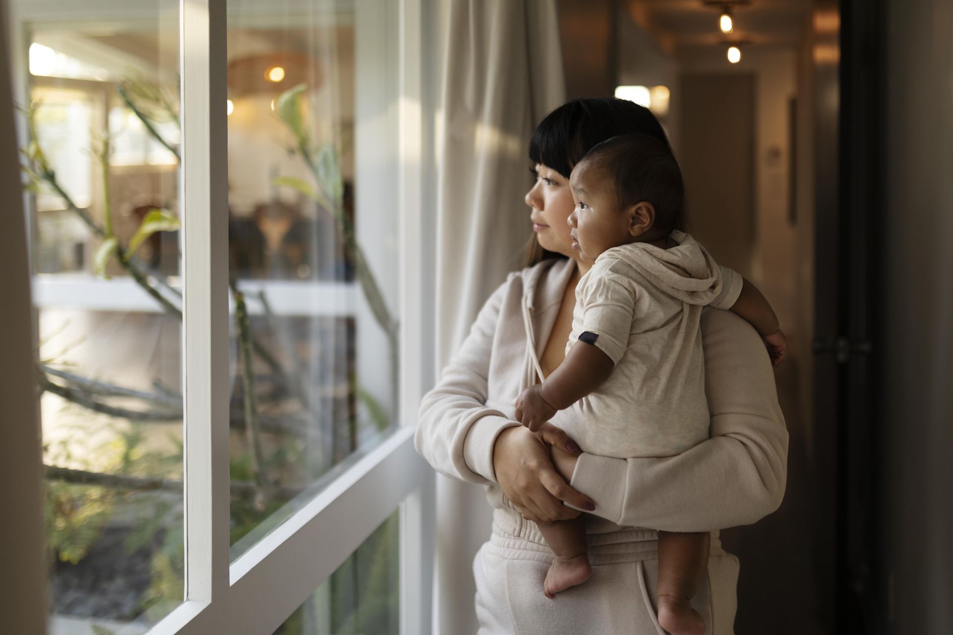 A woman holds a baby while looking out a window in a cozy, softly lit room.