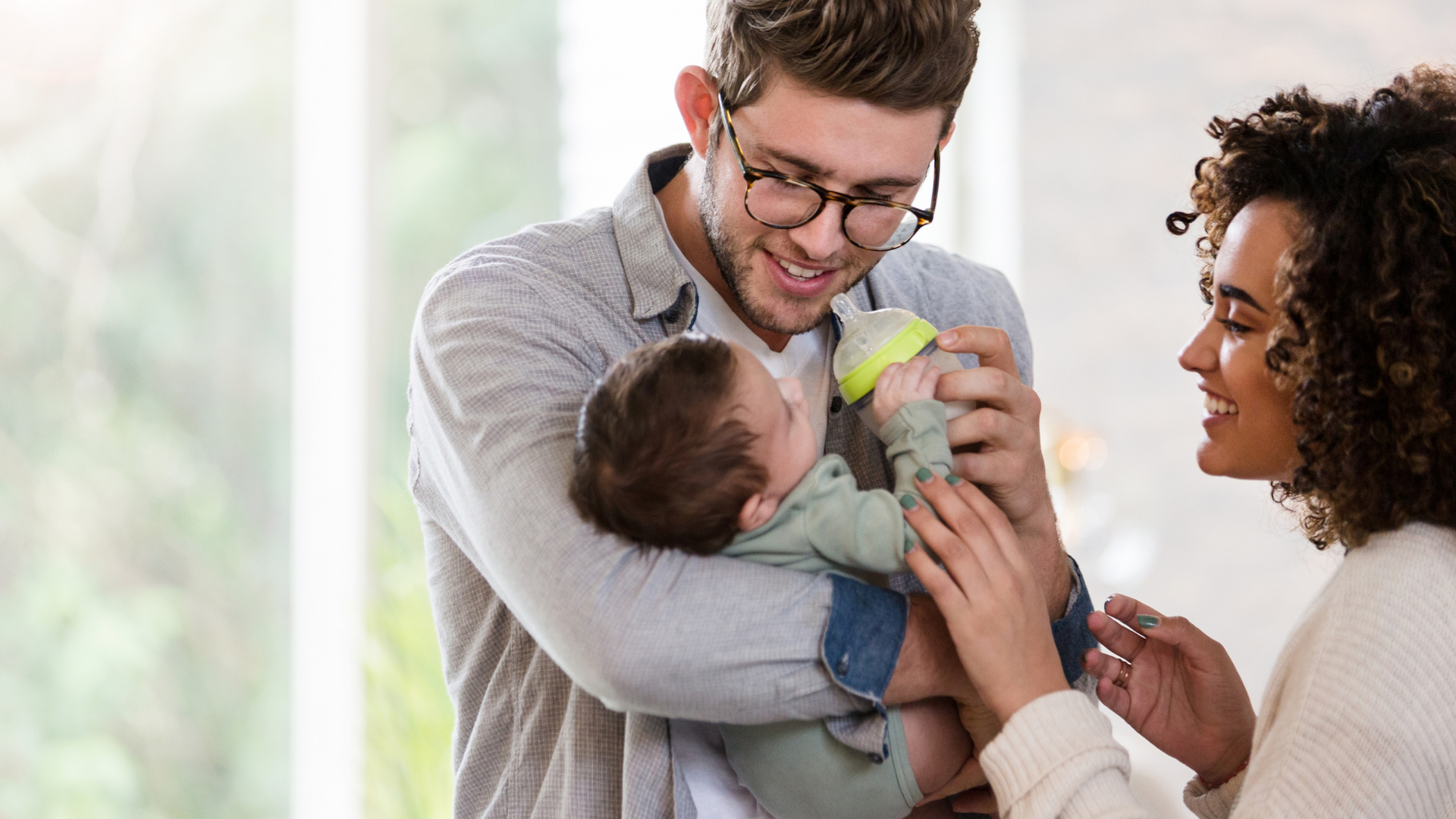 a man feeding a newborn a bottle while a woman watches