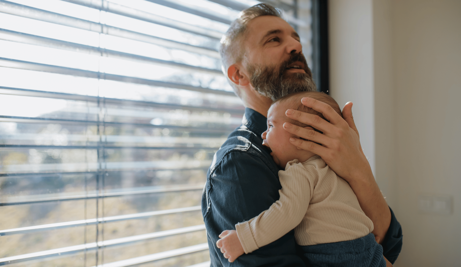 A bearded man holds a newborn by a window, appearing thoughtful.