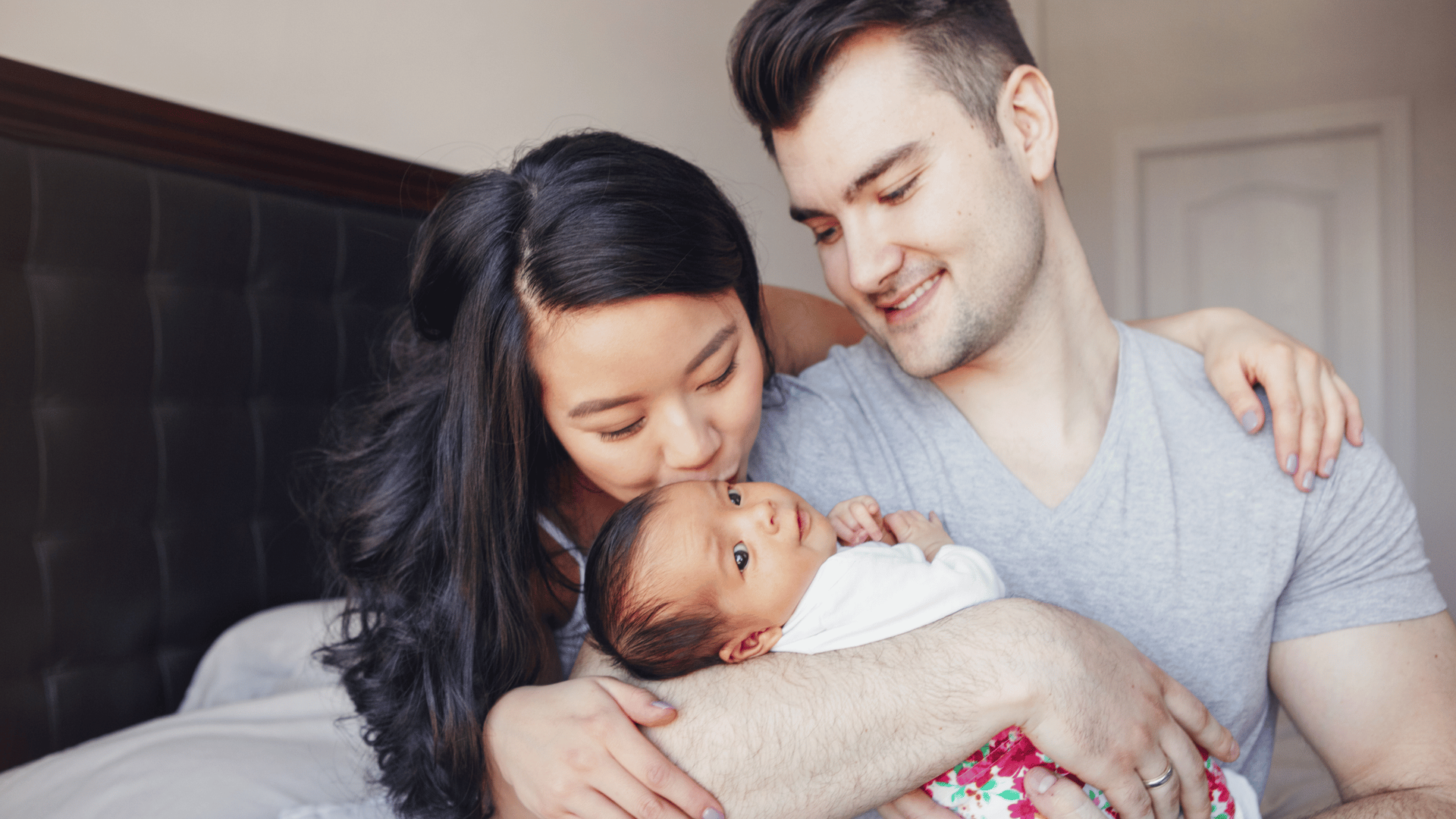 A couple sits closely, warmly embracing their newborn wrapped in a blanket.