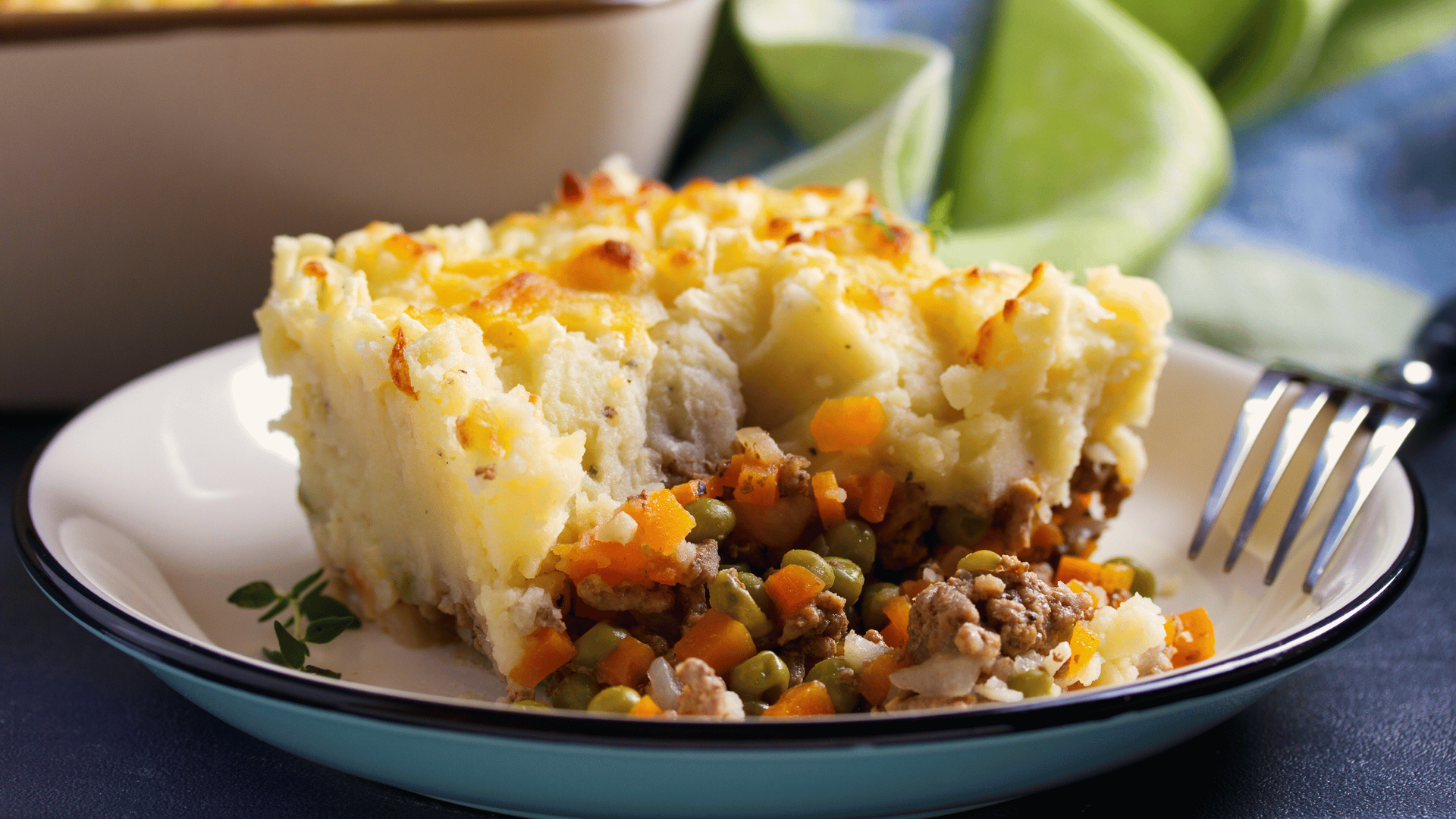 A slice of shepherd's pie on a white plate, featuring creamy mashed potatoes atop minced meat, peas.