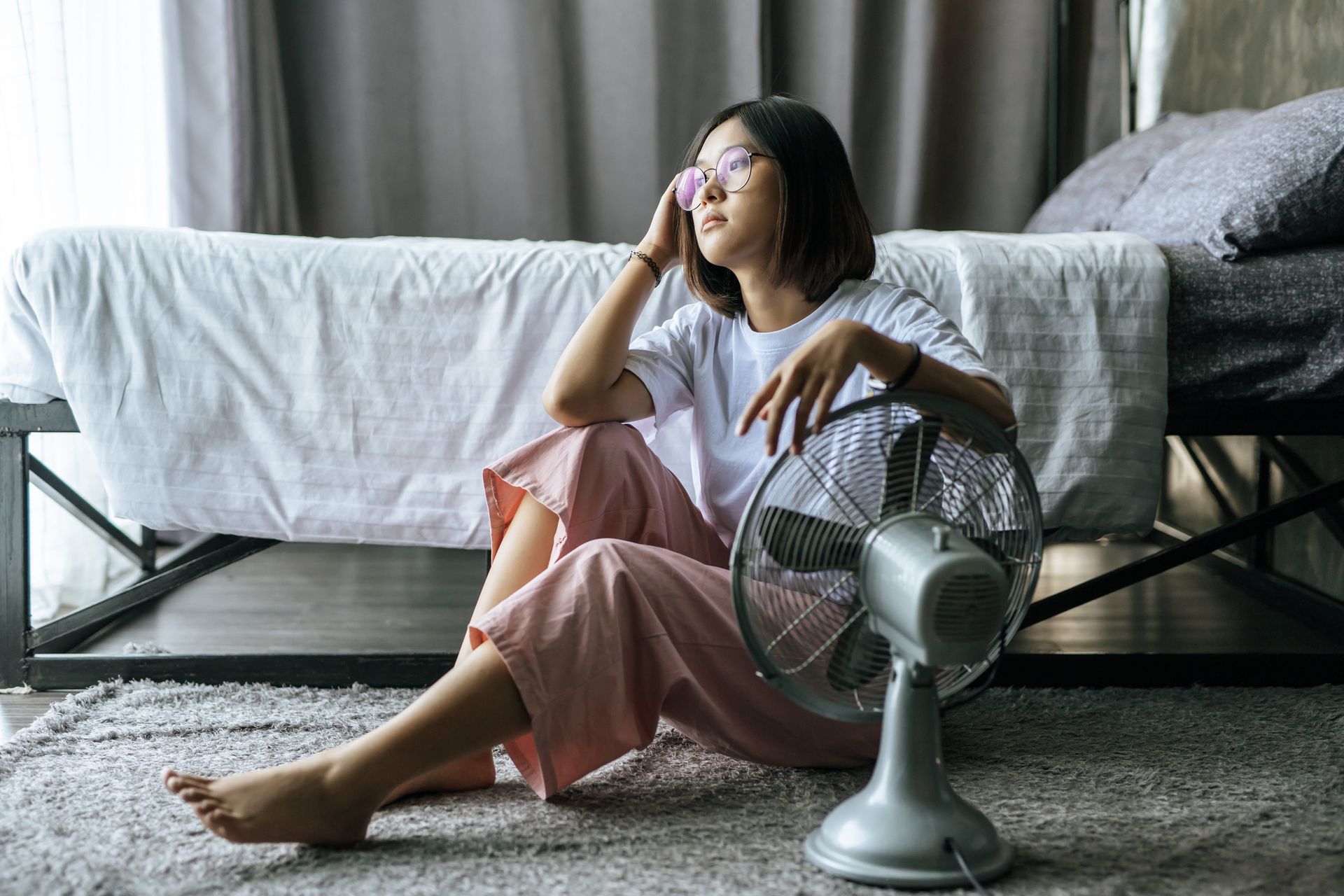 Young woman sitting on the floor beside a bed, with a fan in front of her.
