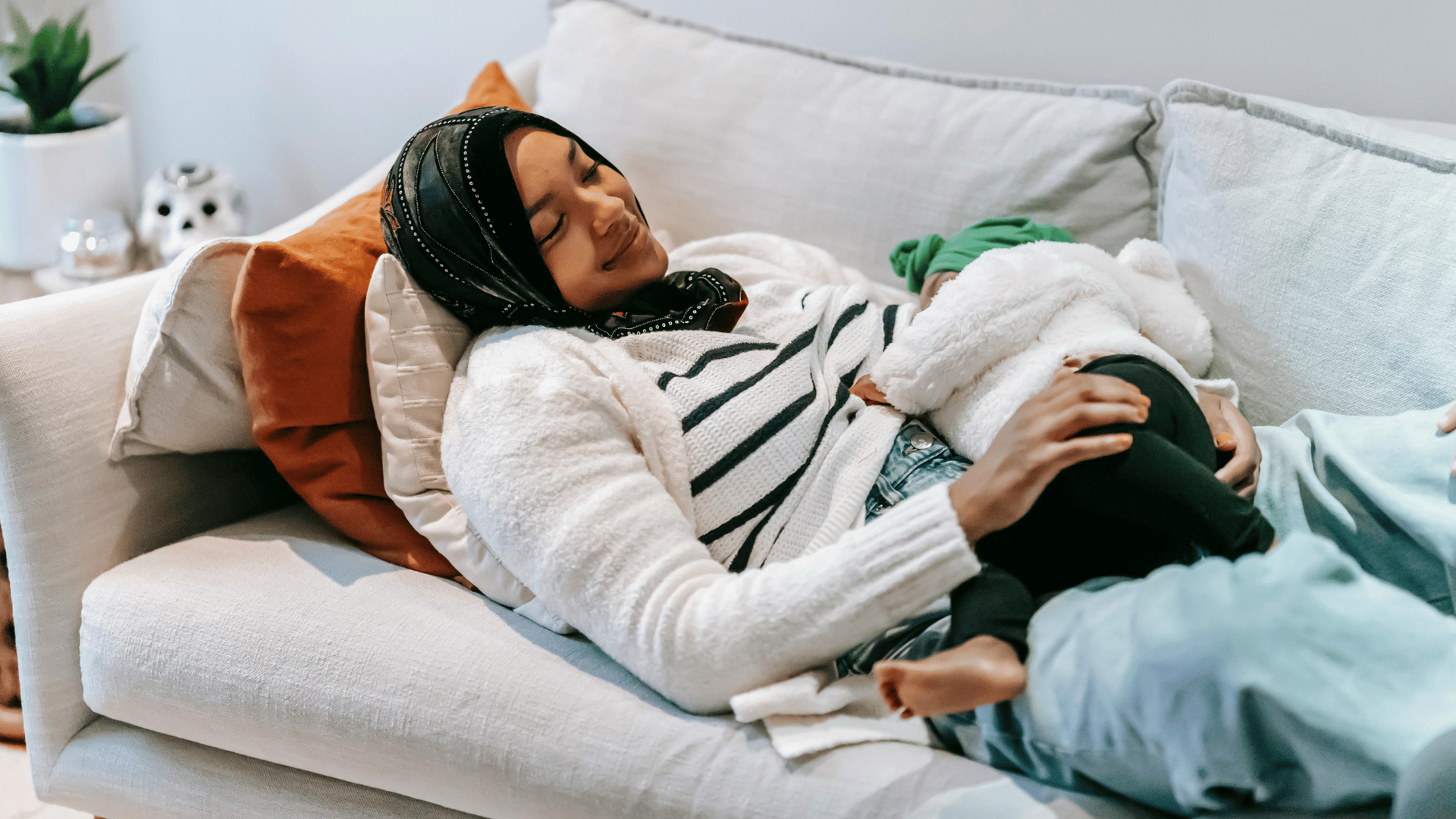 A woman wearing a hijab, striped shirt, and cardigan lays on a white sofa, breastfeeding her baby.