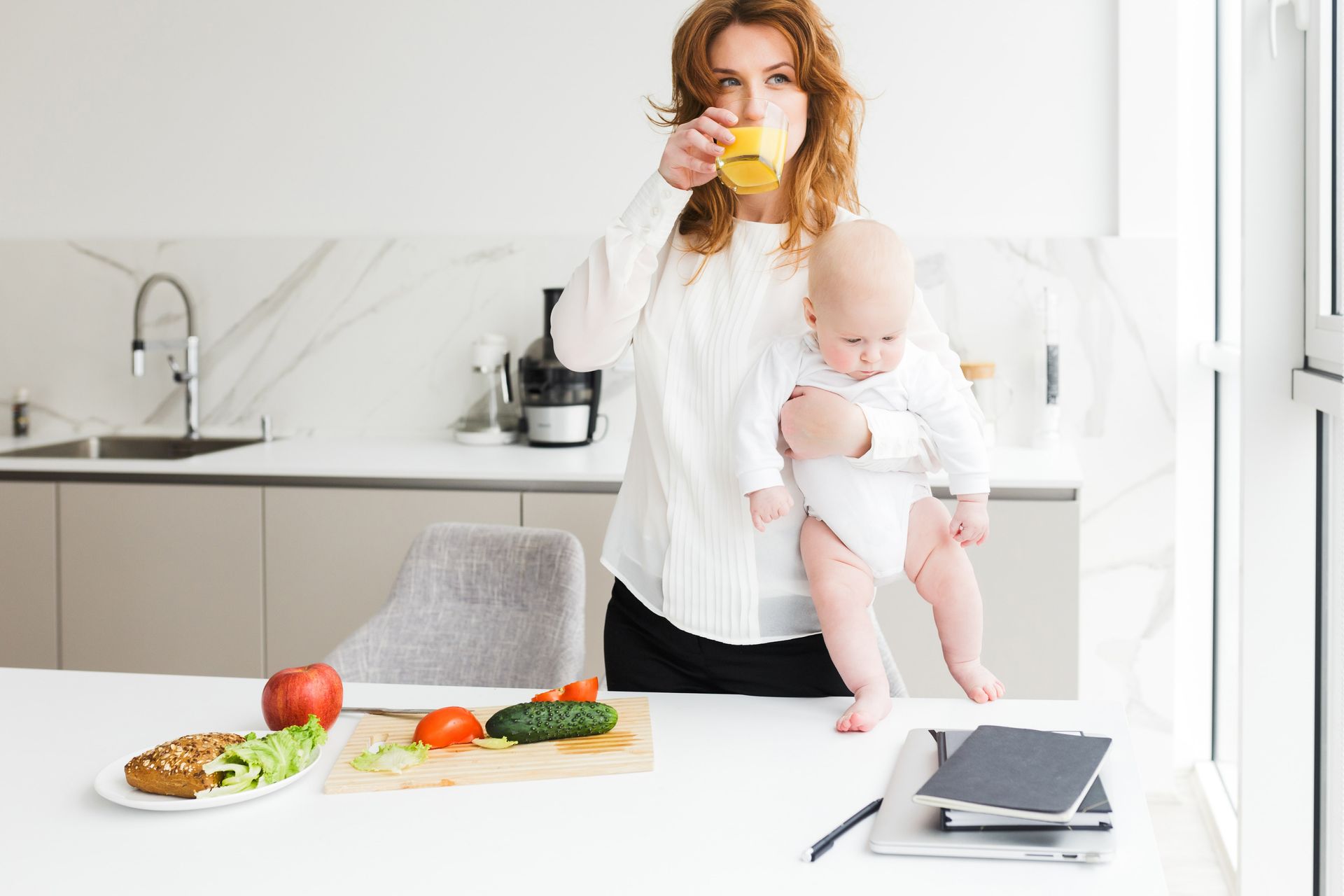 a woman drinking juice while holding her baby with vegetables on the counter