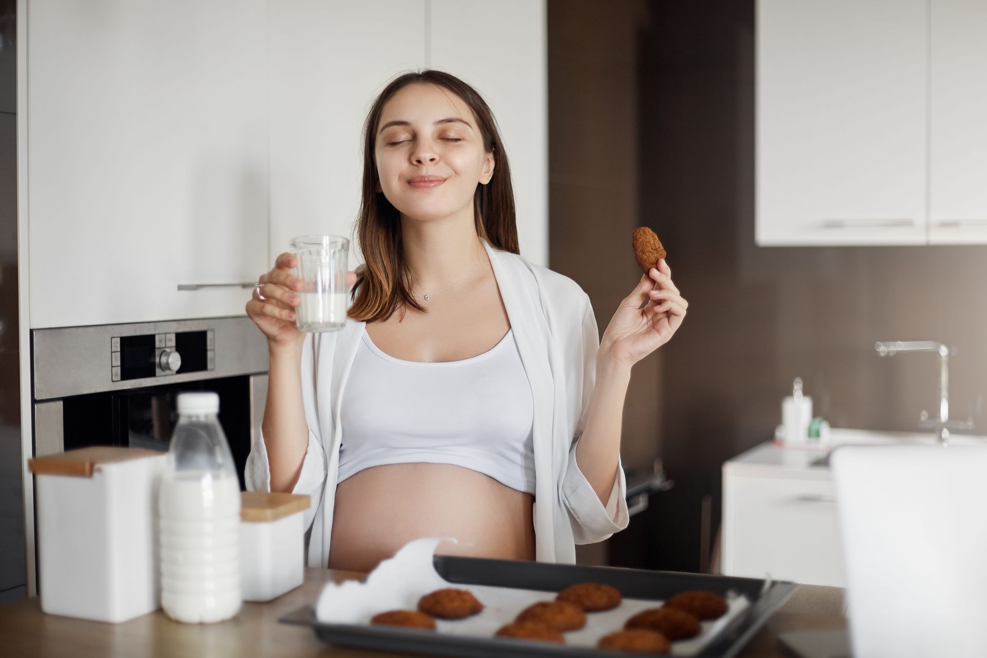 A pregnant woman in a kitchen, smiling, enjoying a cookie and holding a glass of milk.