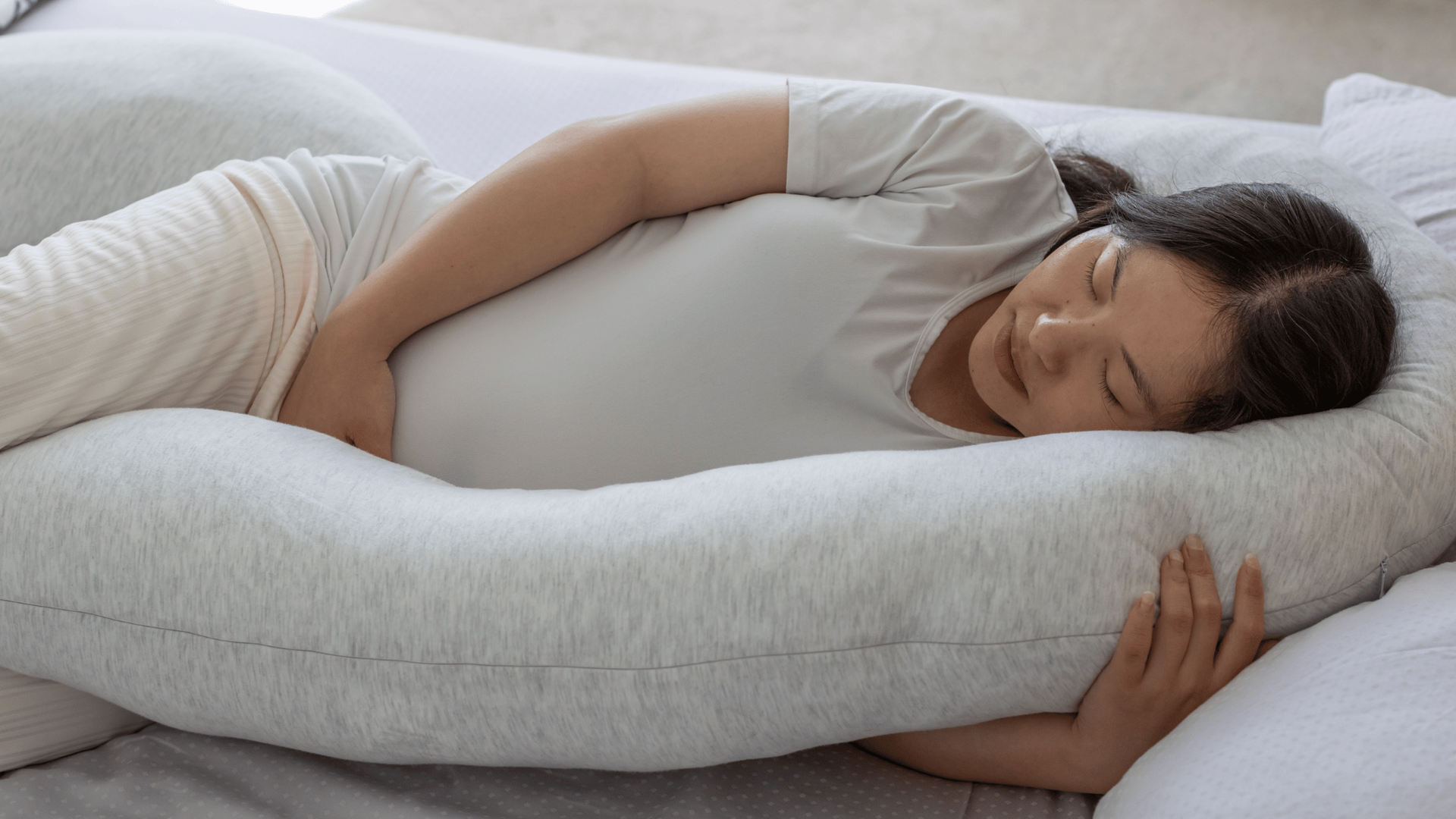 Pregnant woman resting on a large, gray pregnancy pillow, lying on her side in a peaceful bedroom.