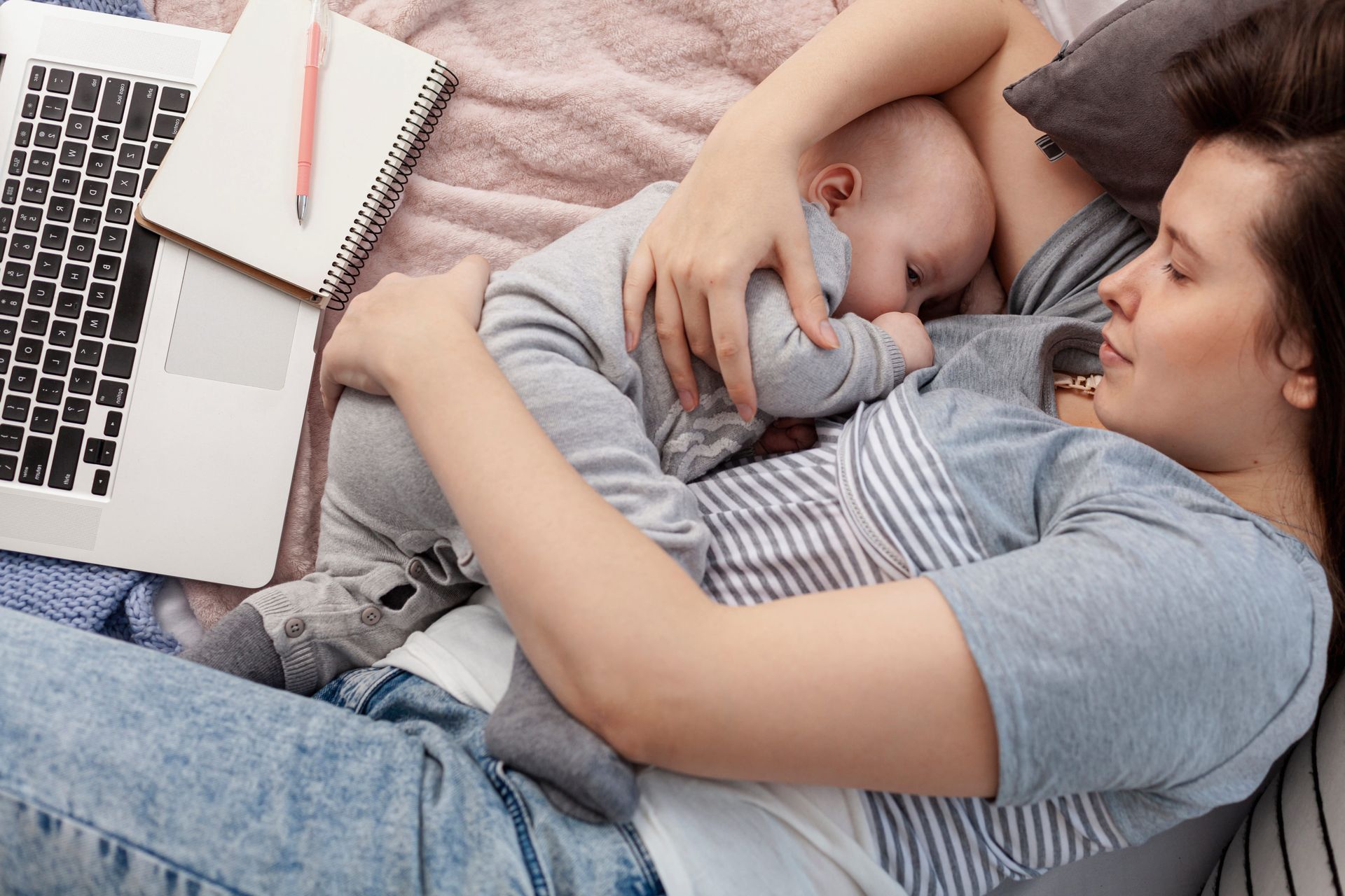 Mother lying on bed, nursing her baby, surrounded by a laptop and notebook with a pen.