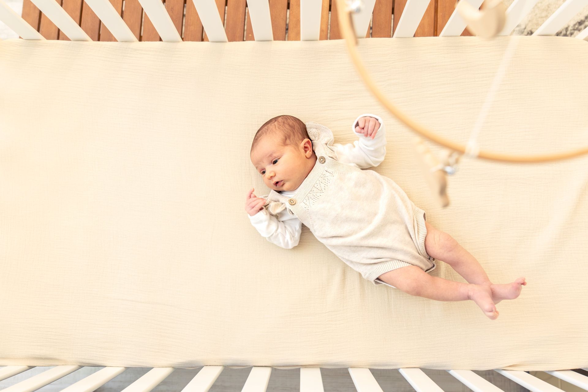 A newborn baby lies in a crib on a soft beige mattress, wearing a cream-colored outfit.
