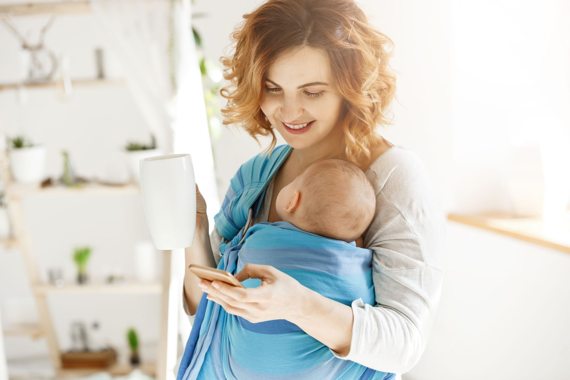 A mother holding her baby and looking at her phone while smiling.