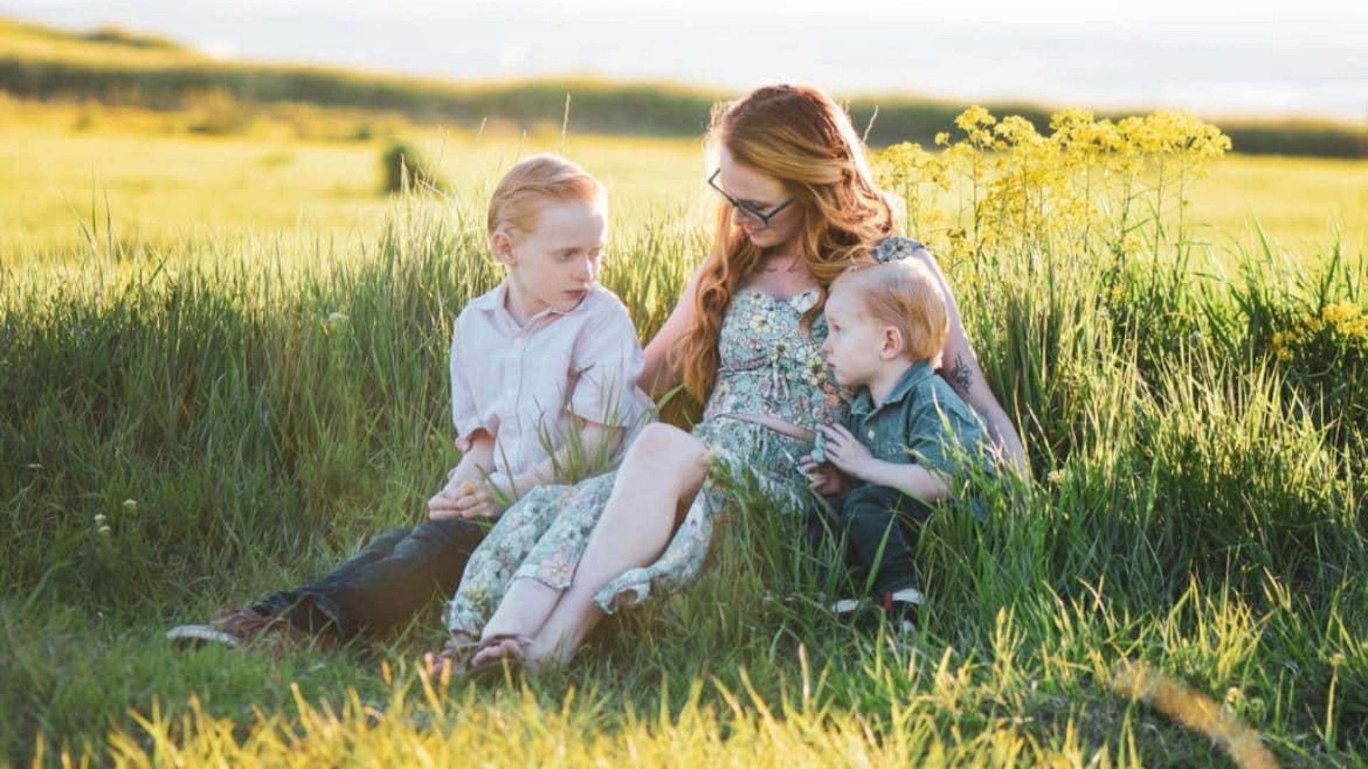 Tiara Monson sits in a field with two children, surrounded by green grass.