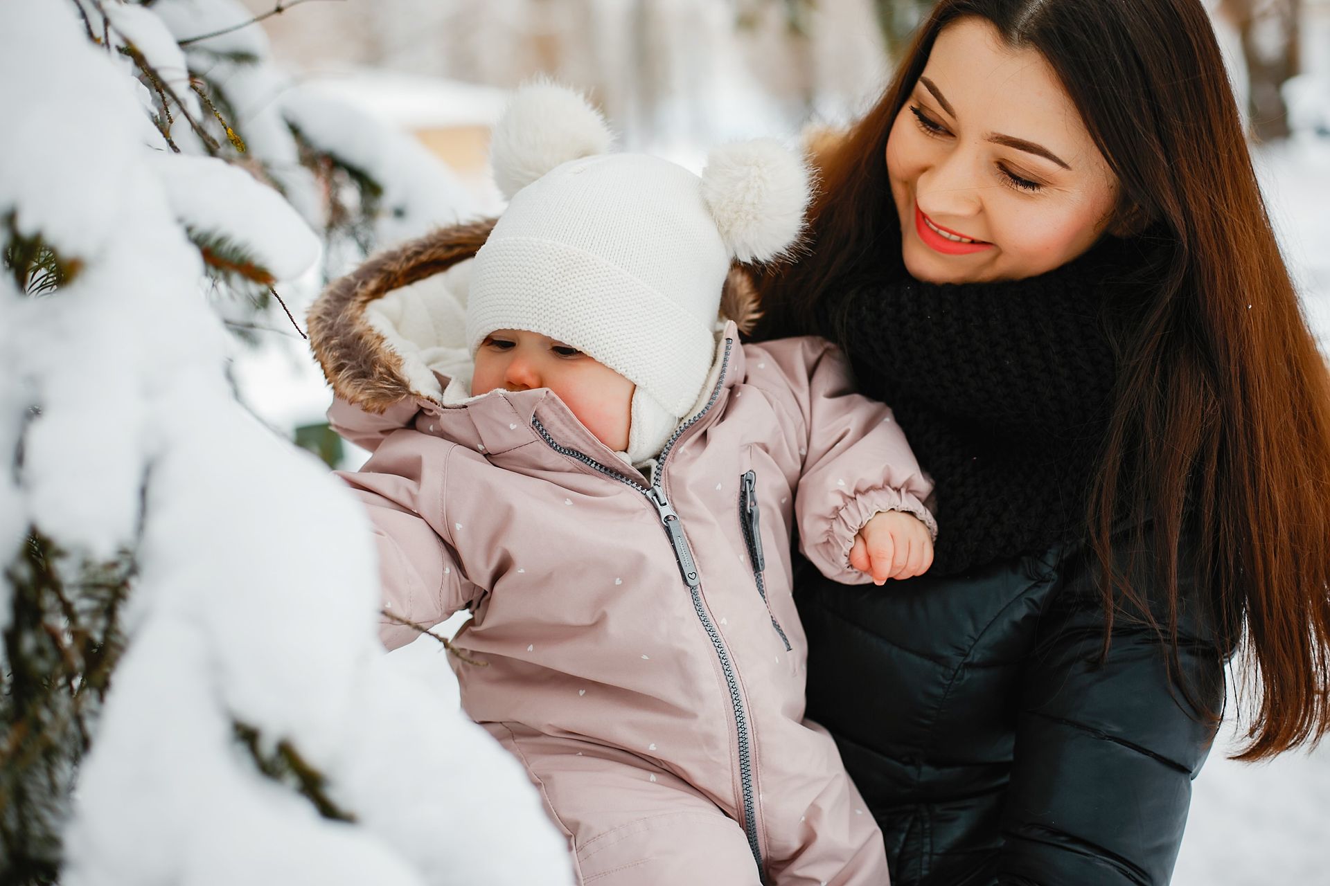 A smiling woman in a black coat holds a baby dressed in a pink snowsuit and white pom-pom hat.
