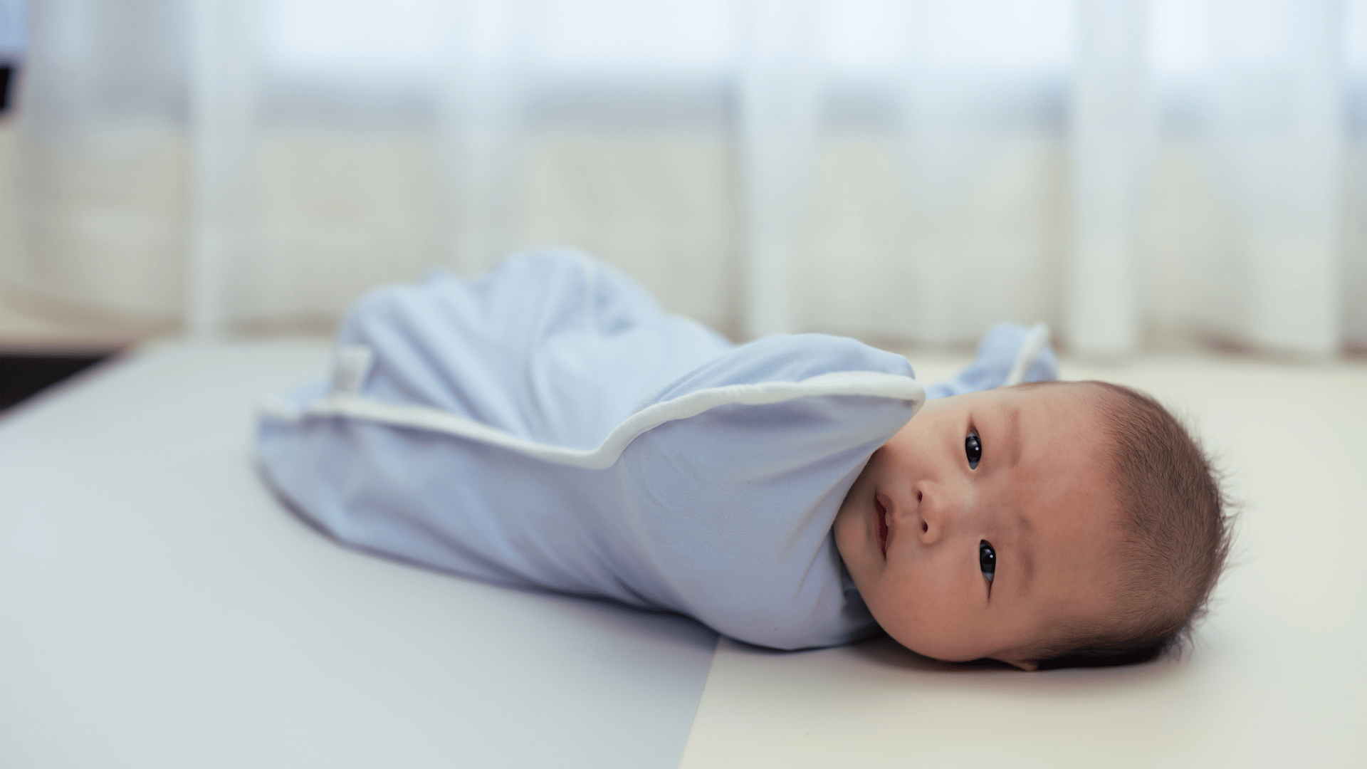 A baby is wrapped snugly in a light blue swaddle, lying on a soft surface.
