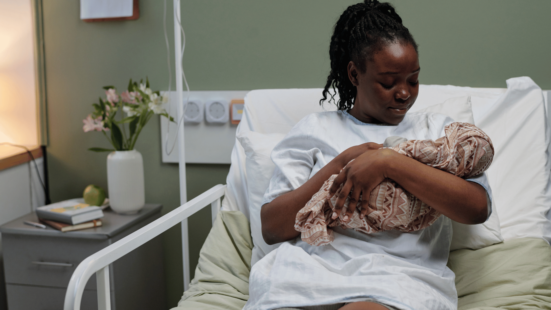 A mother in a hospital gown holds her swaddled baby while sitting in a hospital bed.