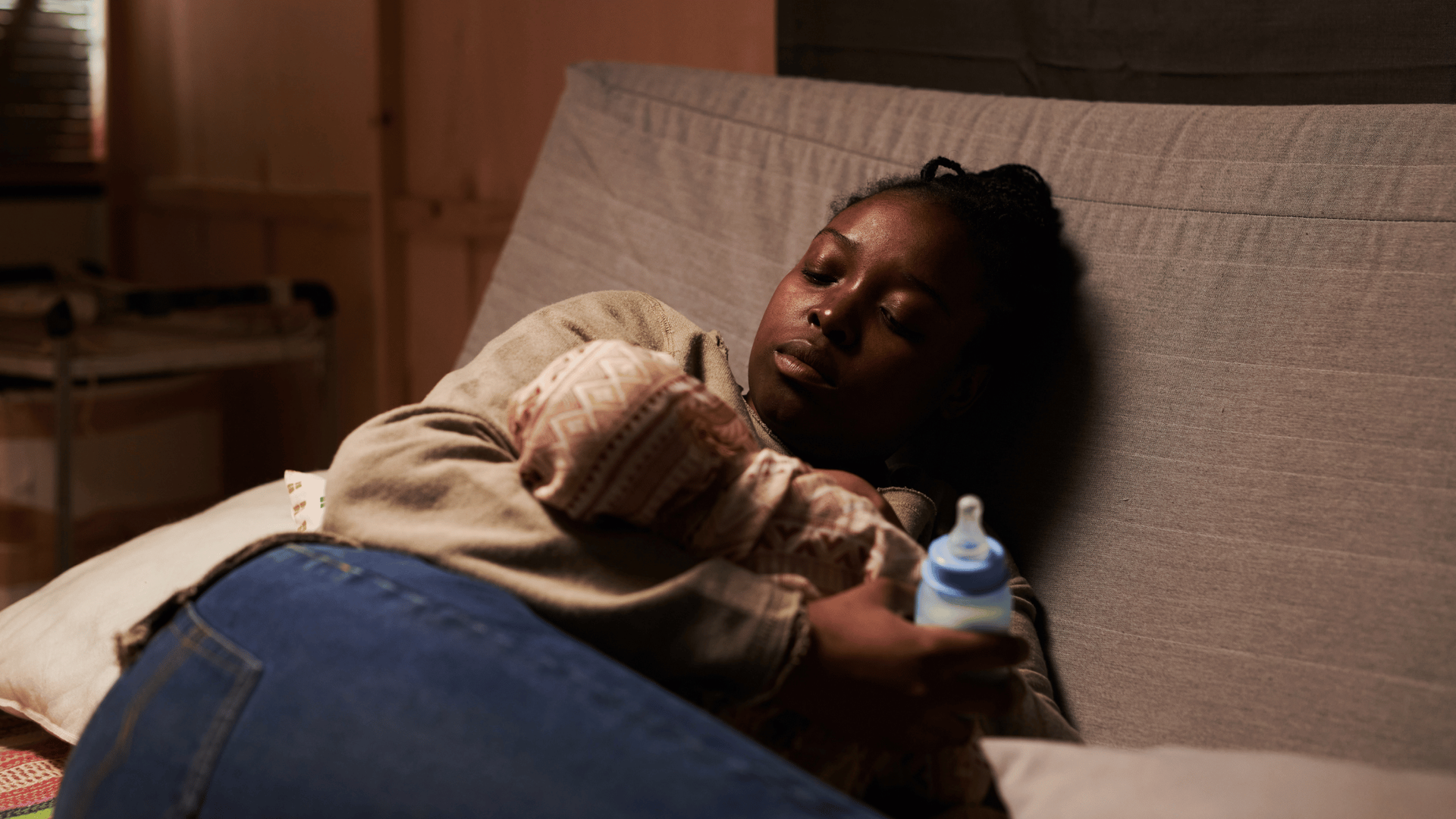 A woman reclines on a sofa, gently holding a baby wrapped in a patterned blanket.