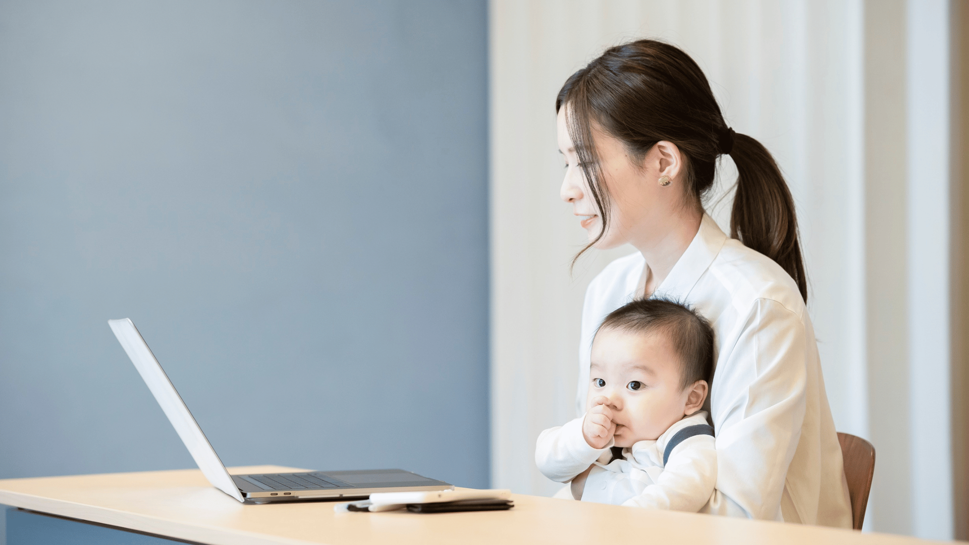 Woman working at a laptop, holding a baby on her lap. The baby is sucking their thumb.