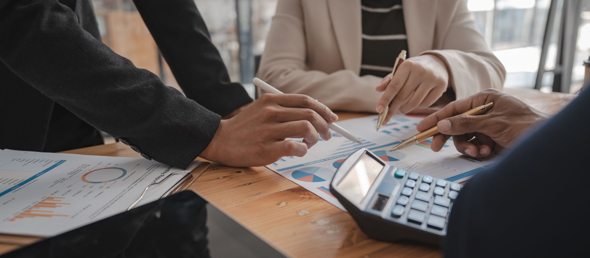 Three accountants reviewing financial charts and graphs with pens and a calculator at a wood desk Three accountants reviewing financial charts and graphs with pens and a calculator at a wood desk