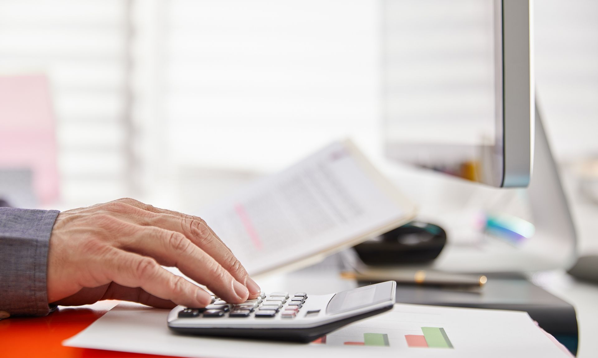 Person's hand using calculator on a desk, near computer screen and documents.
