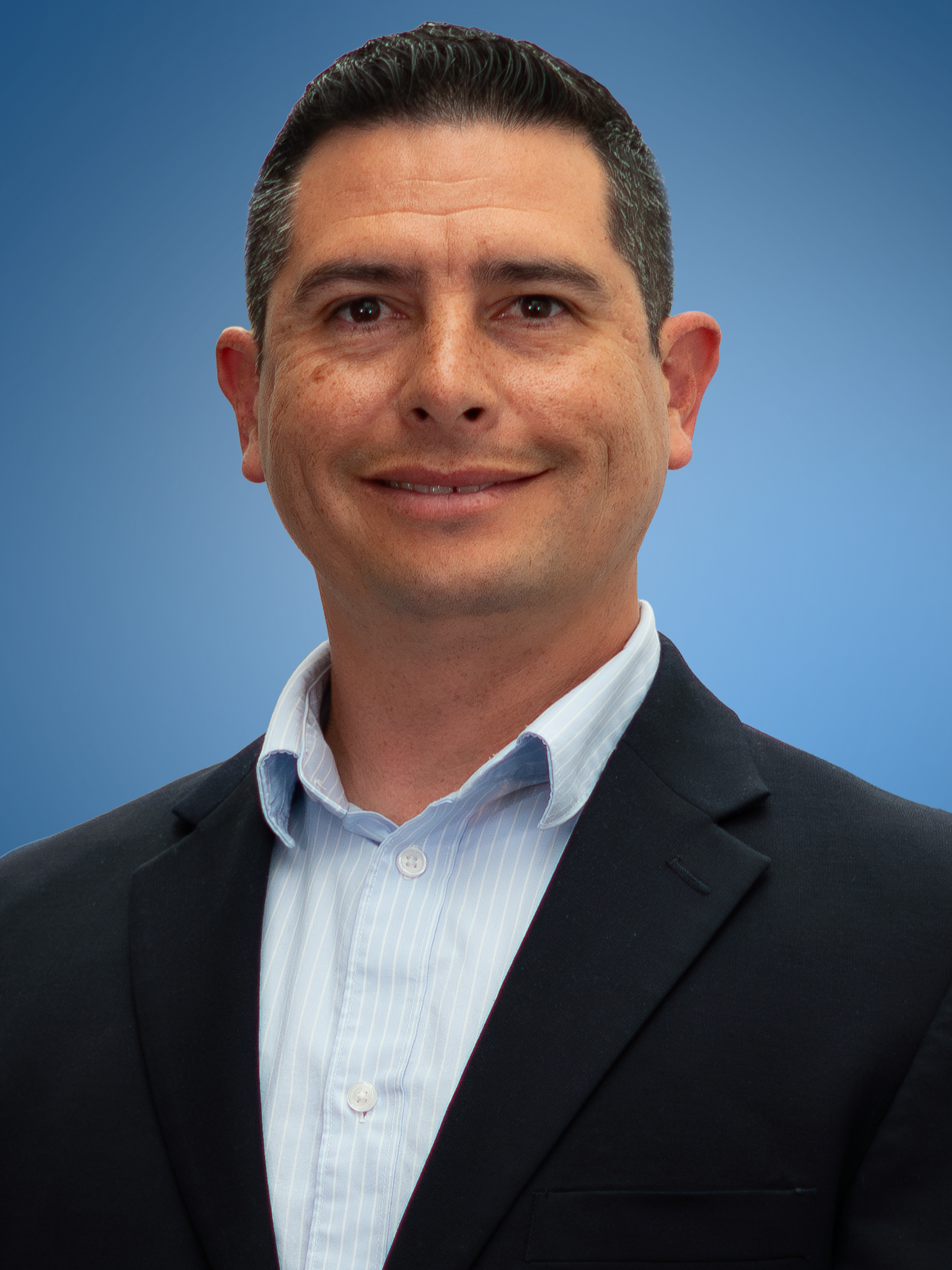 Man in blue shirt, black suit, and striped tie smiles at the camera against a neutral background.