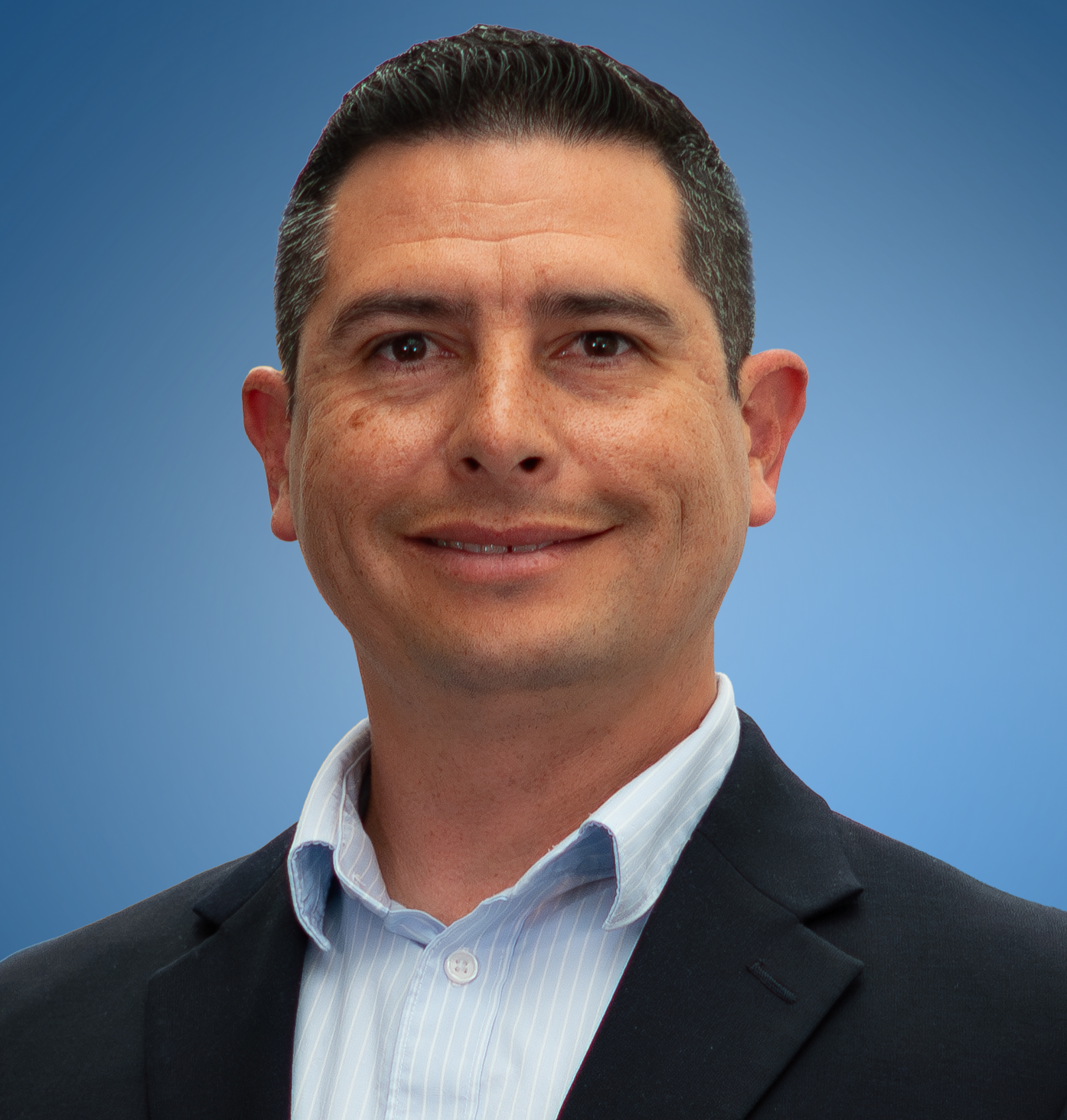 Man in a blue shirt, tie, and black suit, smiling at the camera against a neutral background.