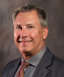 Man in a suit smiles, gray hair, wearing a patterned tie, against a blurred background.