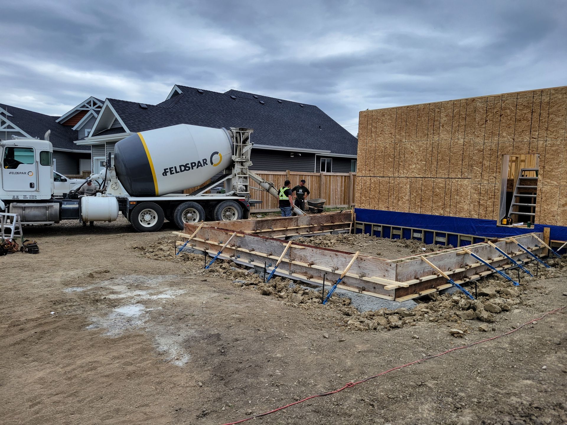A concrete mixer truck is driving down a dirt road next to a house under construction.