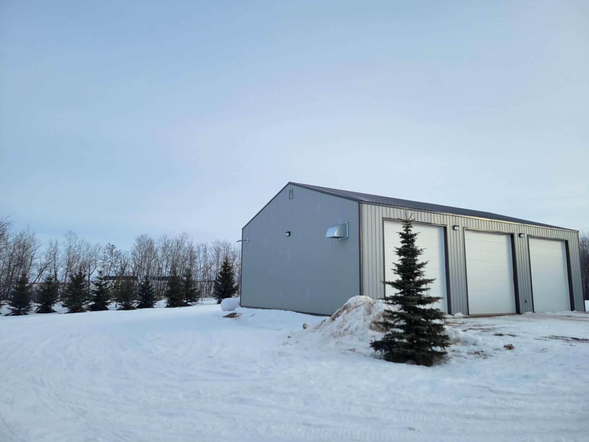 A garage with three doors is surrounded by snow and trees
