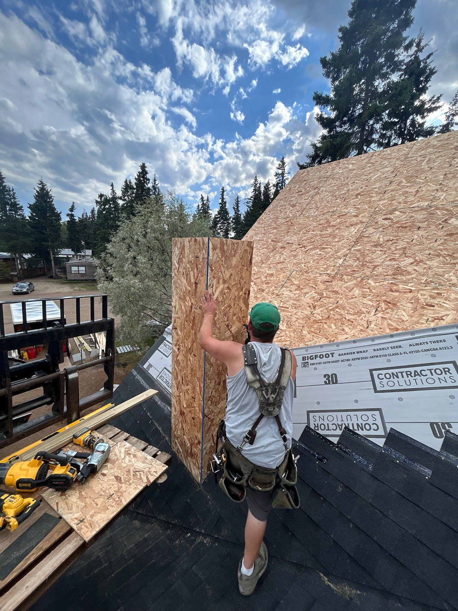 Roofer on a roof installing wood paneling. Cloudy sky, trees in the background, tools on the roof.