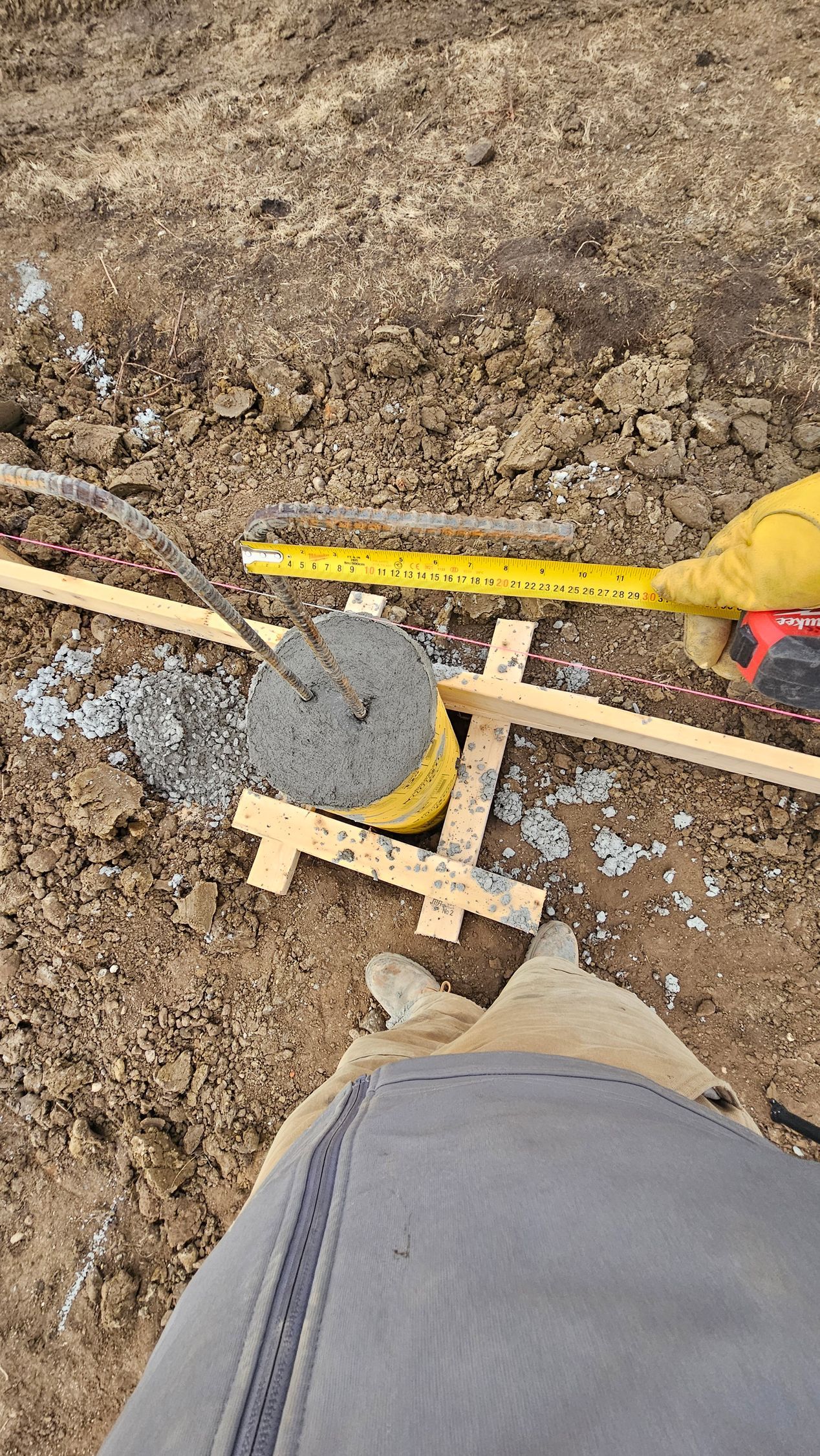Person using a tape measure to check a small concrete pillar in dirt, wooden structure around it.