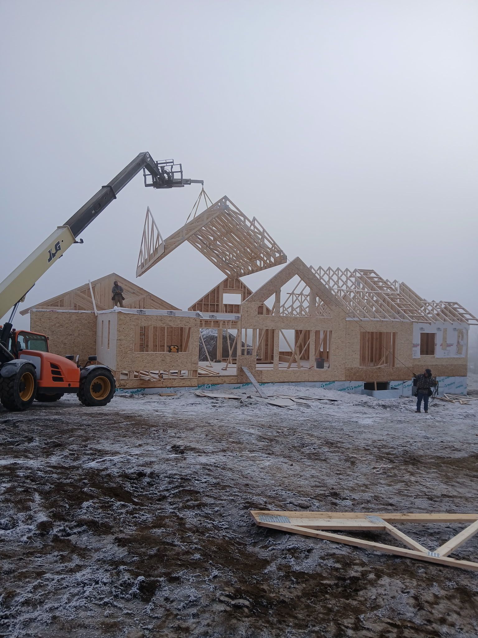 Construction of a wooden house with a crane lifting a roof truss in a snowy field.