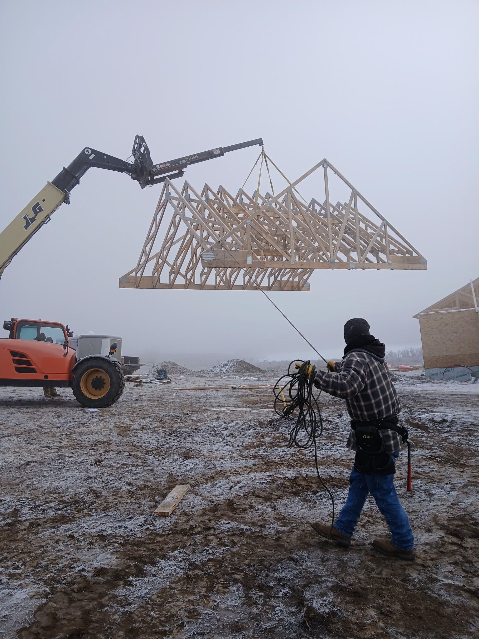 A telehandler lifts a wooden roof truss. A construction worker steadies it on a foggy day.