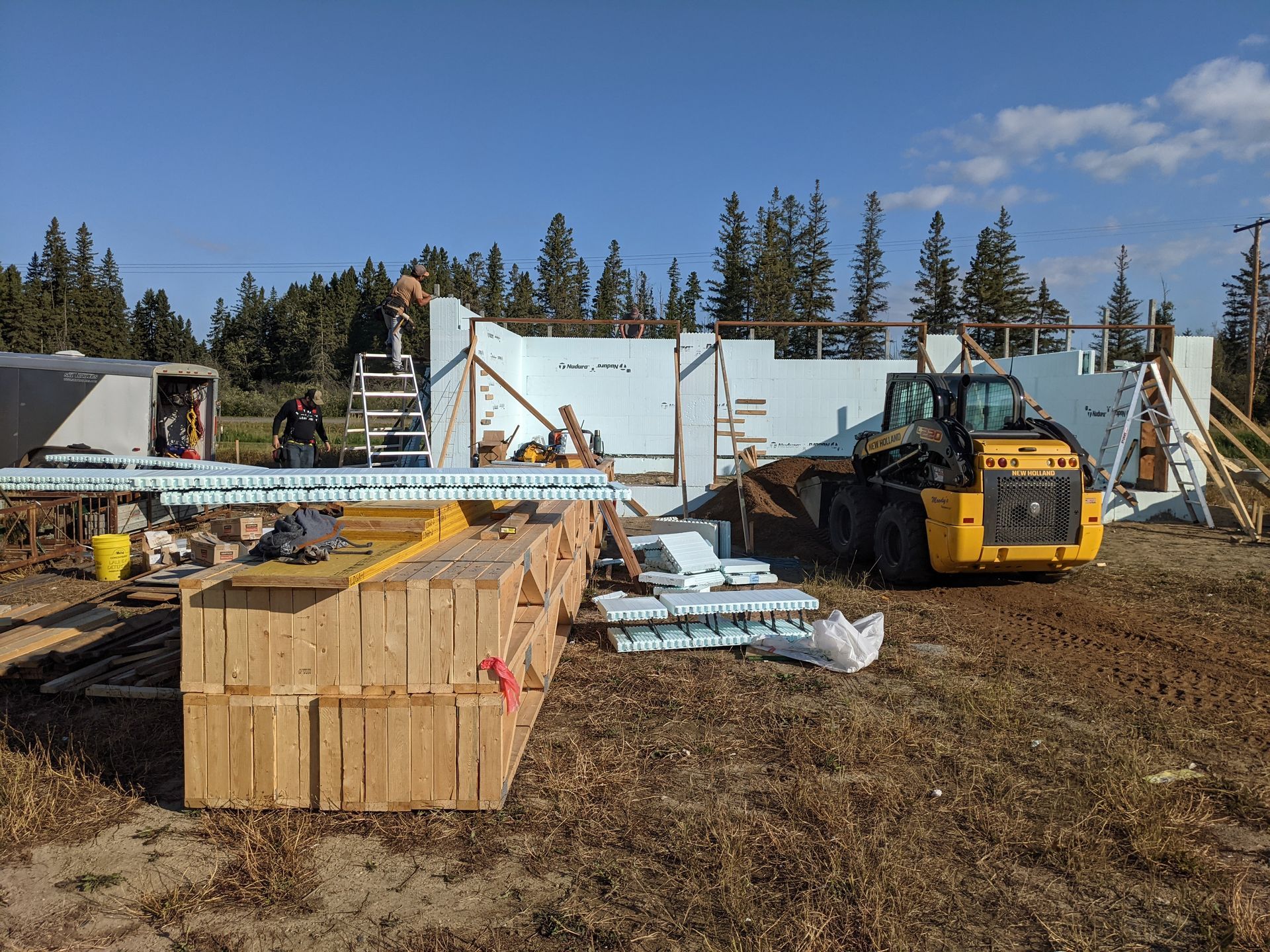 Construction site with a partially built white insulated concrete form (ICF) structure, with a yellow skid steer loader nearby.