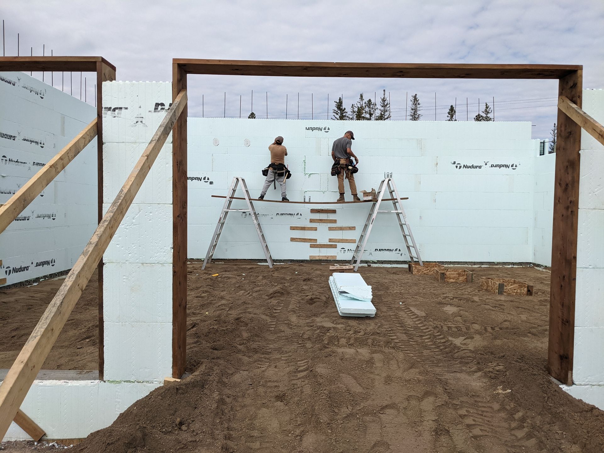 Two construction workers on ladders inside a structure with foam walls. They work on a wall with wooden frames.